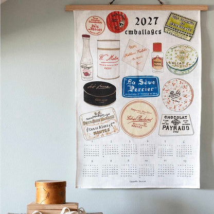 A wooden-framed wall calendar hanging on a light blue wall, featuring vintage French candy and cosmetic packaging illustrations with the text &