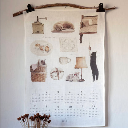 A fabric wall calendar hanging from a rustic wooden rod with black clips, featuring vintage-style illustrations of cats, a coffee grinder, a lamp, and a calendar grid for 2027. The calendar is displayed against a plain off-white wall with dried flowers at the bottom.