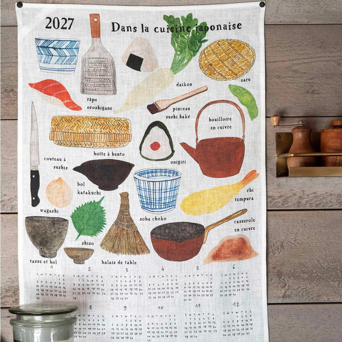 A vertical fabric wall calendar hanging on a rustic wood wall, featuring illustrated Japanese foods and utensils with French labels and a 2027 calendar grid at the bottom.