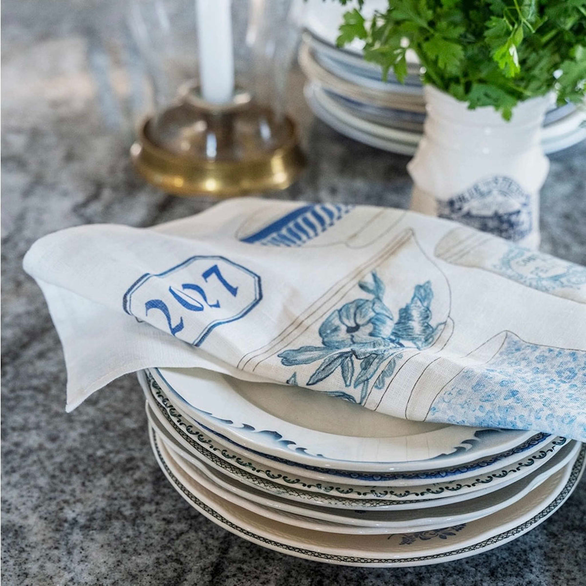 A stack of white ceramic plates with blue patterns and a matching white linen cloth with blue floral and architectural illustrations are arranged on a gray stone countertop. The dishcloth has the number 2027 printed on it.