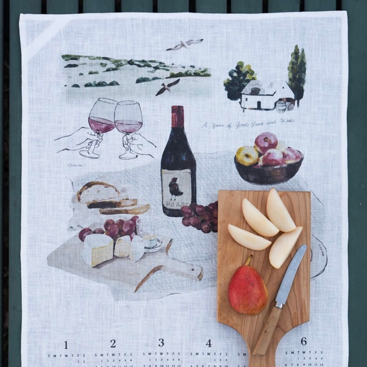 A white linen tea towel with a wine-themed kitchen print, laid flat on a dark green slatted surface. The print features illustrations of wine glasses, a wine bottle, grapes, cheese, bread, and a wooden cutting board with sliced pears and a knife.