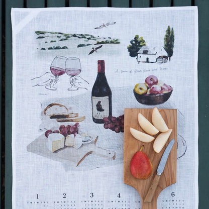 A white linen tea towel with a wine-themed kitchen print, laid flat on a dark green slatted surface. The print features illustrations of wine glasses, a wine bottle, grapes, cheese, bread, and a wooden cutting board with sliced pears and a knife.