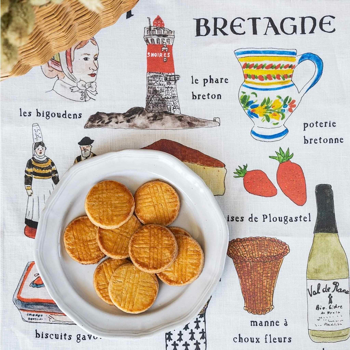 A white ceramic plate filled with golden-brown French biscuits sits on a printed linen fabric cloth featuring Breton-themed illustrations and text, with a wicker basket edge visible at the top left.