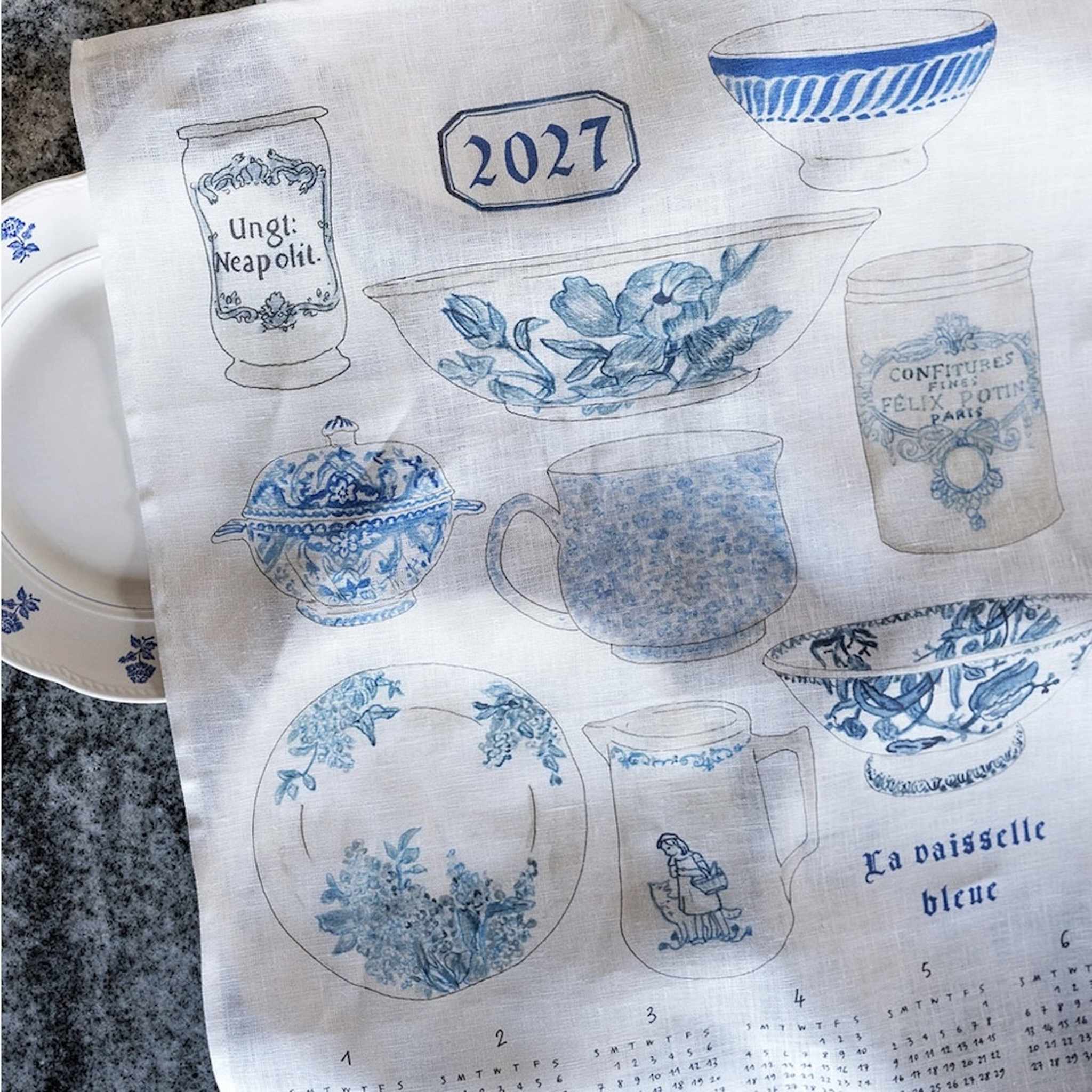 A white linen fabric tea towel printed with blue vintage ceramic motifs, including a teapot, cups, plates, and a calendar at the bottom. It is laid flat on a dark stone surface with partial white dishes visible at the edges.