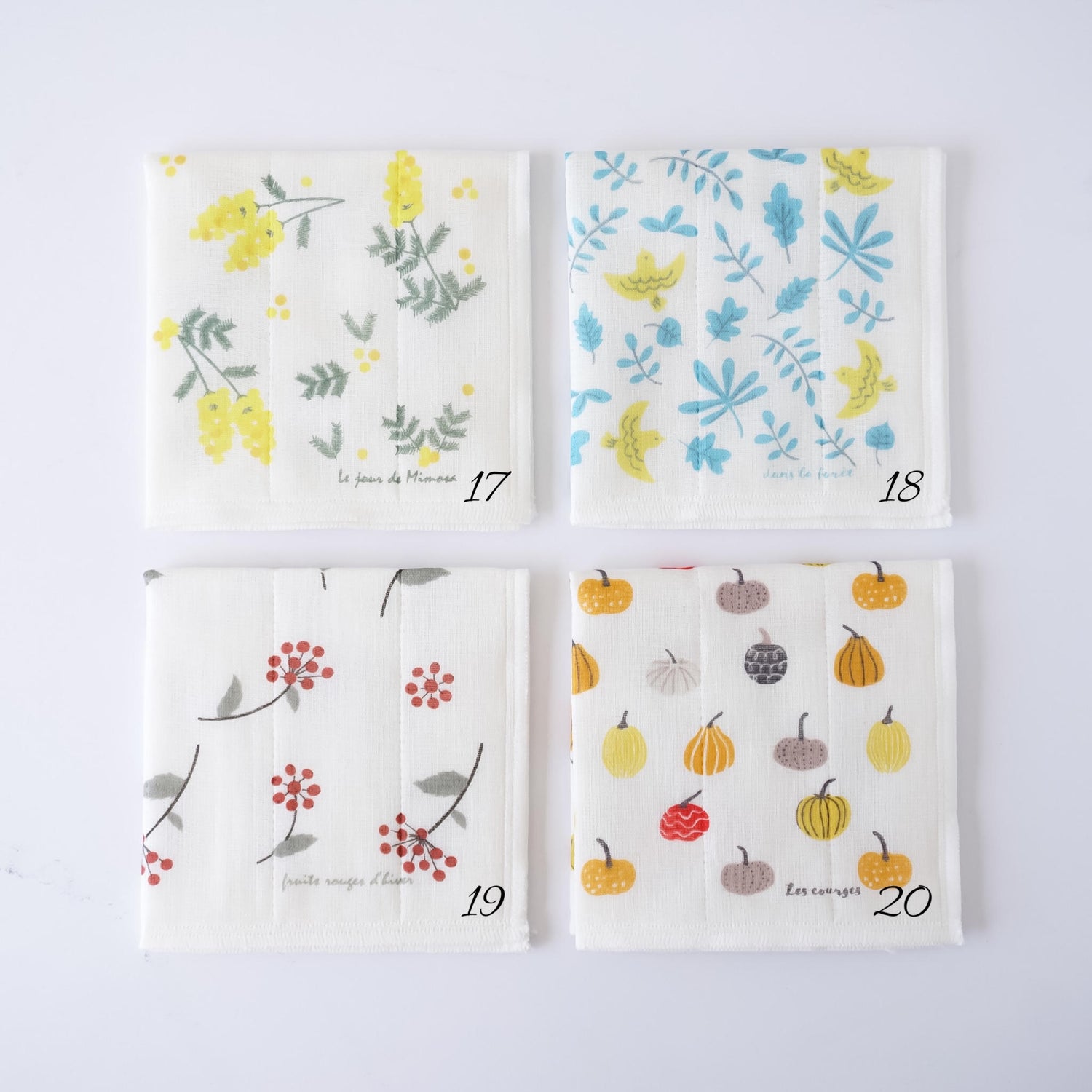 Four square fabric dishcloths with different autumn-themed prints, arranged in a 2x2 grid on a white background. The prints feature yellow flowers, blue leaves, red berries, and pumpkins on an off-white base.