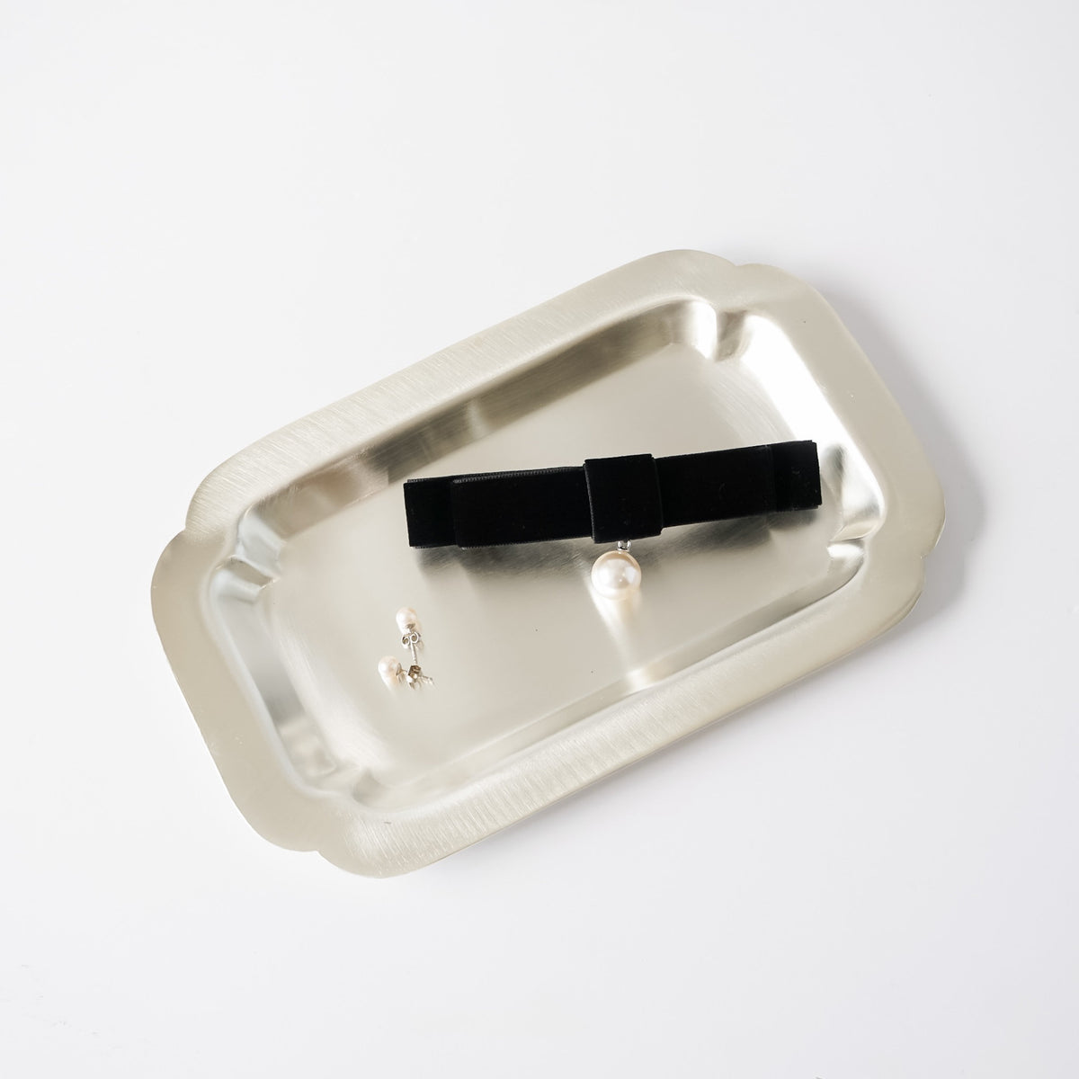 A black velvet bow hair clip with a small pearl accent, displayed on a brushed silver metal tray against a clean white background.