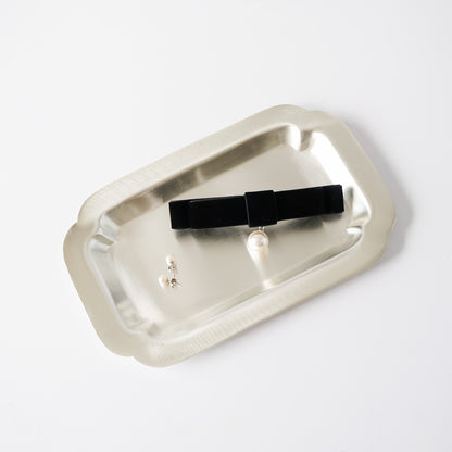 A black velvet bow hair clip with a small pearl accent, displayed on a brushed silver metal tray against a clean white background.
