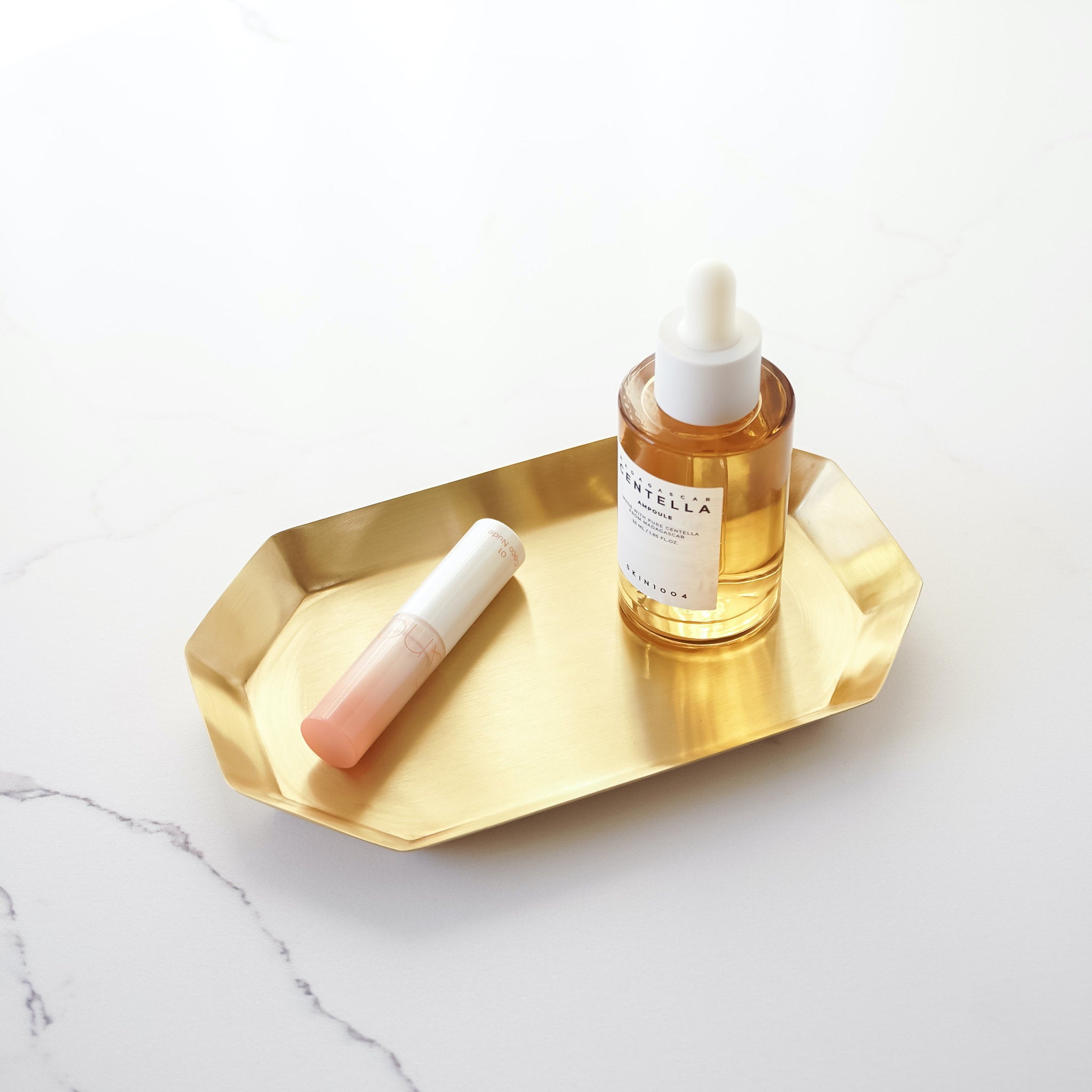 A small, antiqued brass long octagon tray on a marble surface, holding a bottle of liquid and a lipstick.