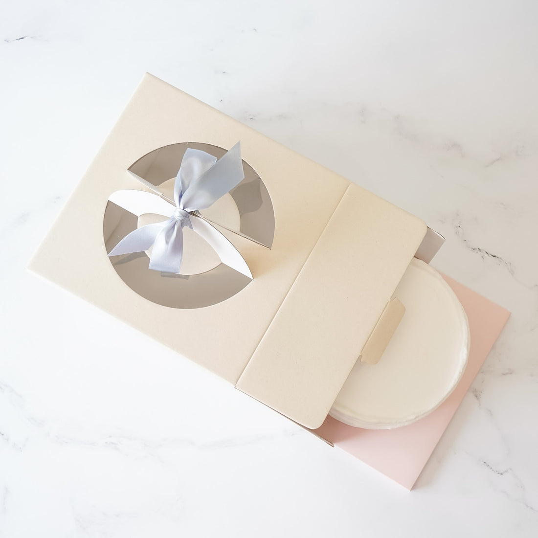 A beige cake box with a clear window displaying a cake, tied with a blue ribbon.