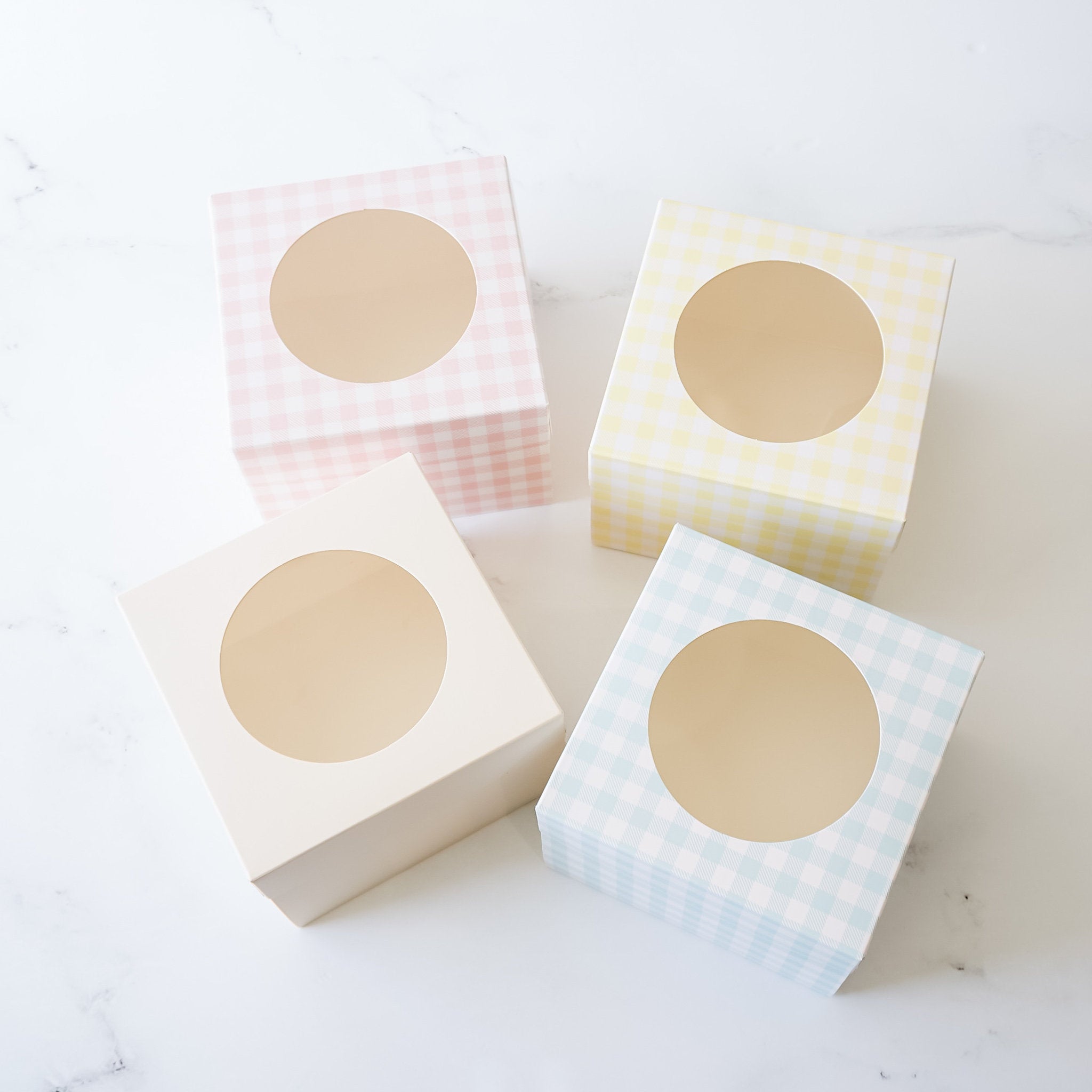 Four square gift boxes with a window. The boxes are made of cardboard and have a checkered pattern. The boxes are in four different colors: pink, yellow, white, and blue.