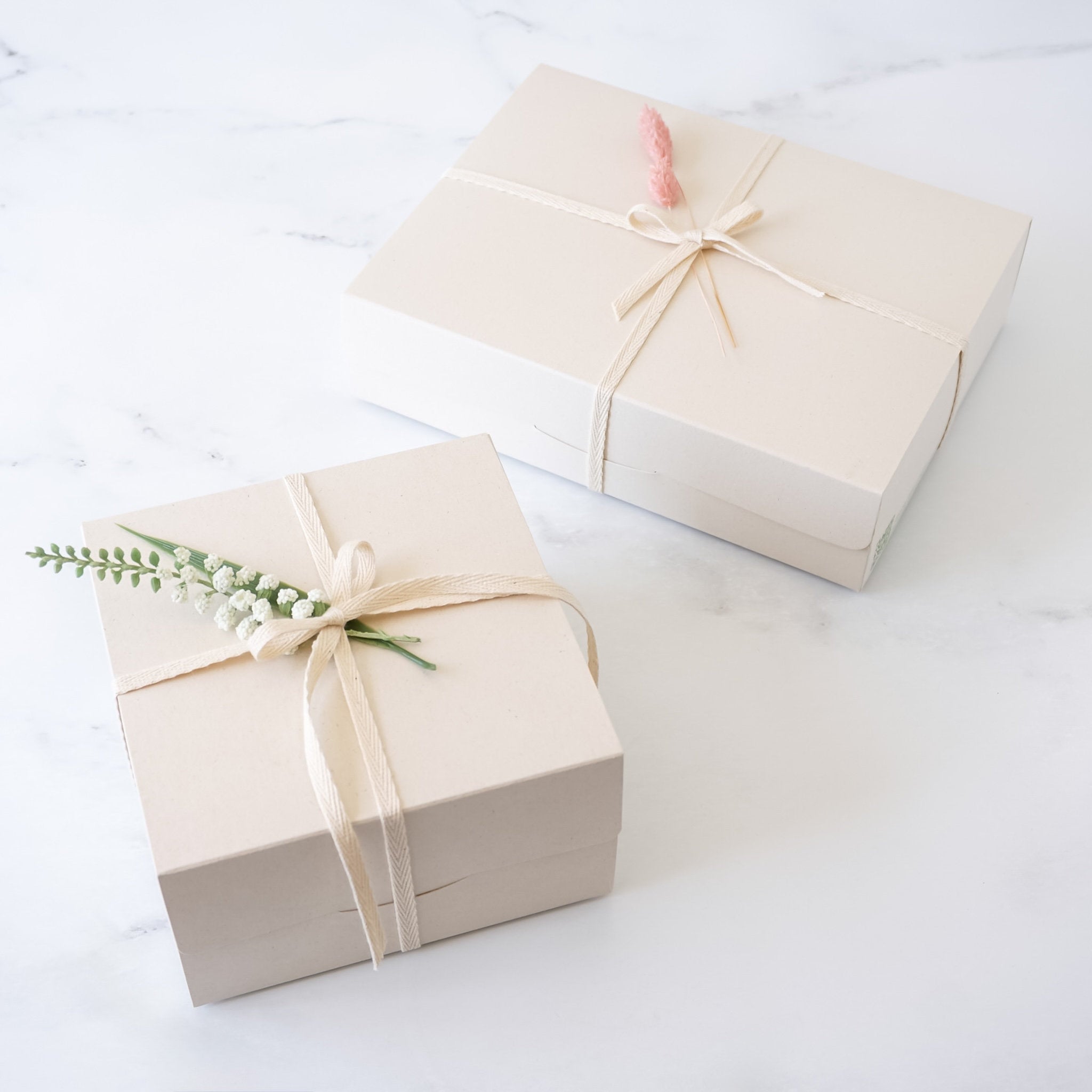 Two light beige bakery boxes with a natural paper texture, adorned with a satin bow and a sprig of dried flowers.