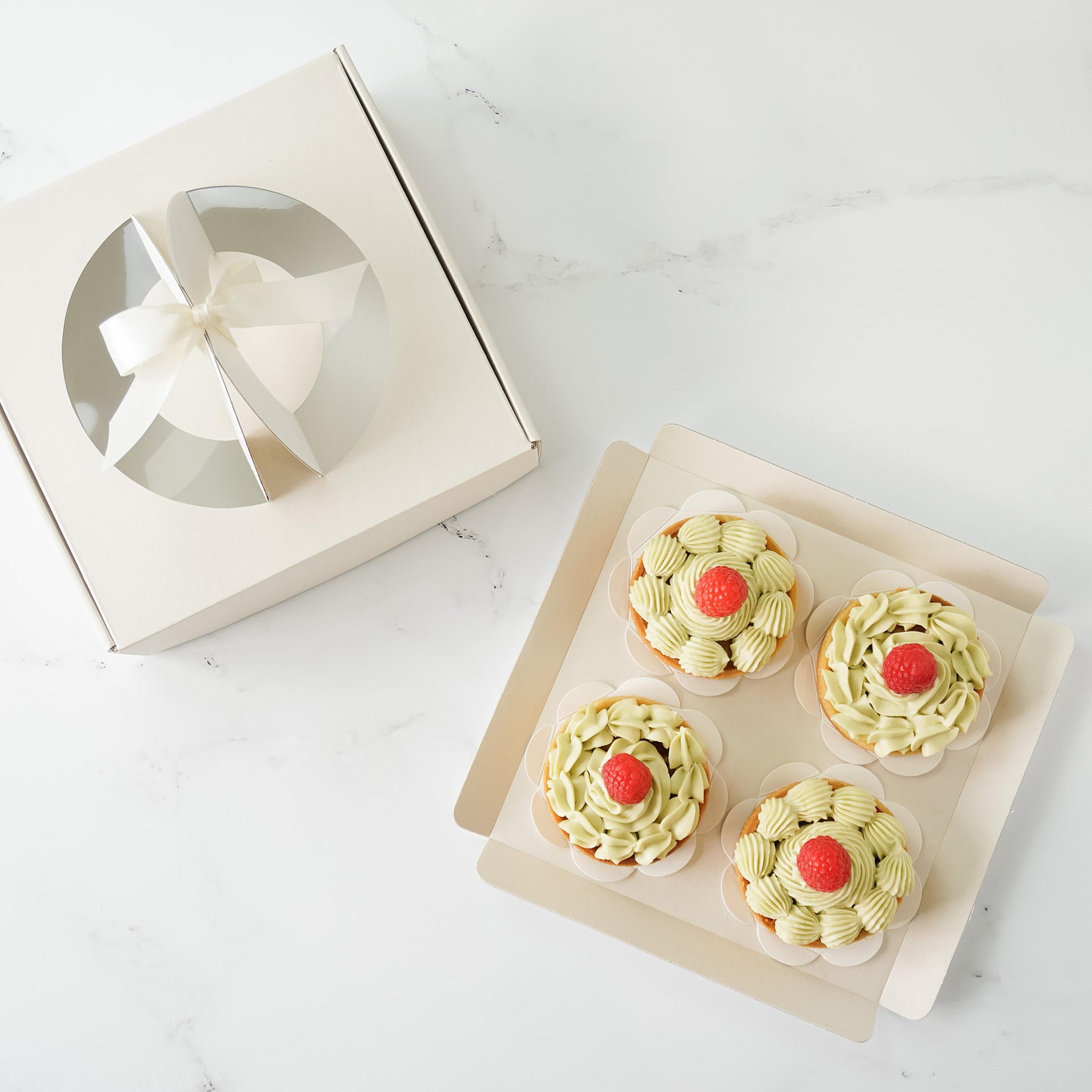 Four green and white frosted cupcakes with a single red raspberry on top, arranged in a white cardboard box.