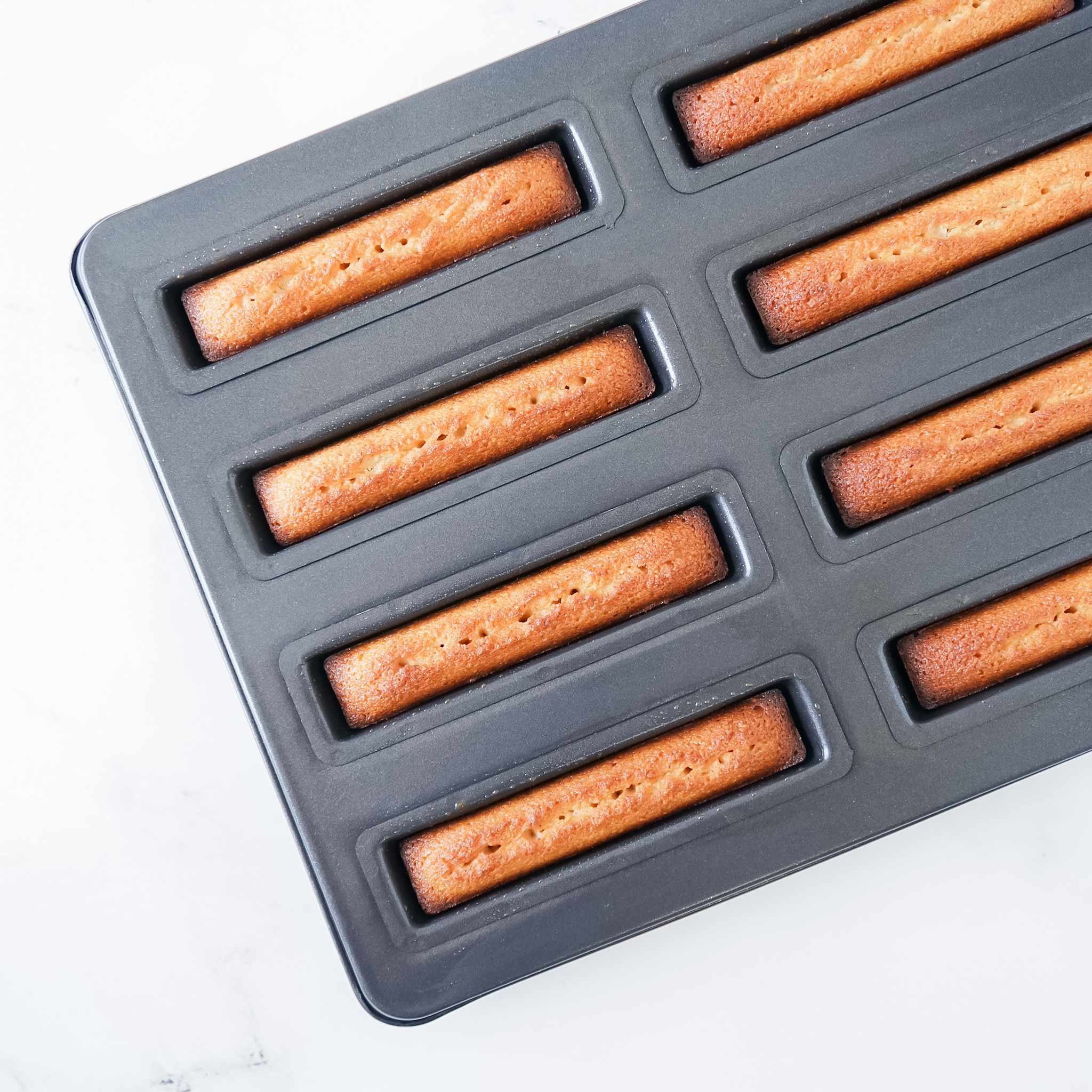 A black financier cake pan with eight cavities, each shaped like a baton. The pan is made of cold-rolled steel with a nonstick coating.