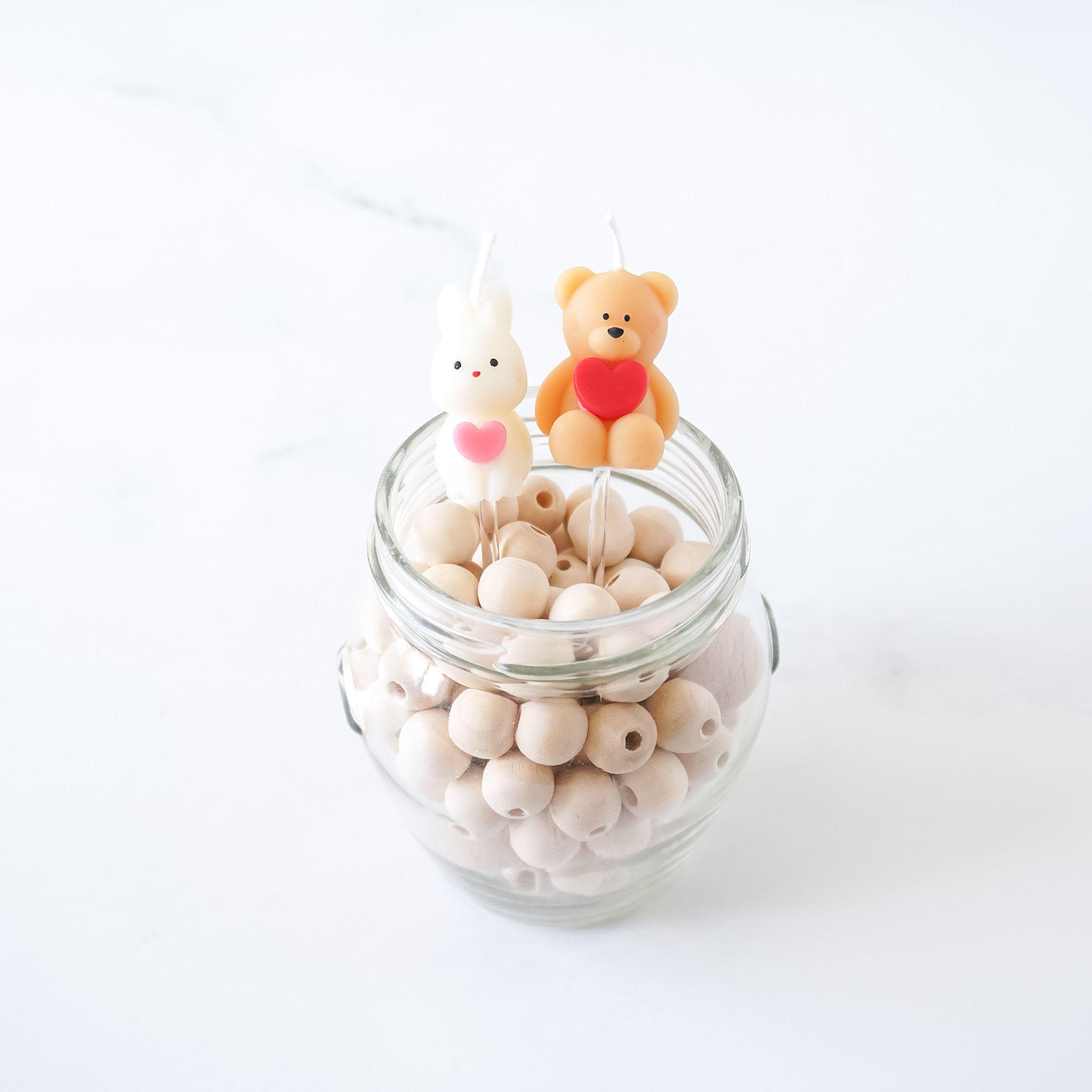 A glass jar filled with white wooden beads with two candles on top. One candle is a white bunny with a pink heart and the other is a brown bear with a red heart.
