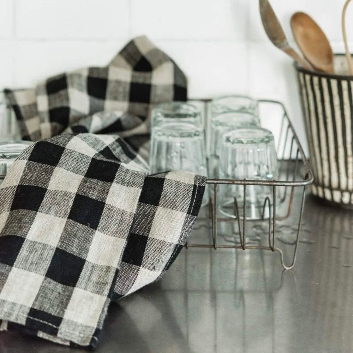 A black and beige buffalo plaid kitchen towel draped over a metal dish rack holding clear drinking glasses on a stainless steel countertop, with a striped utensil holder and wooden spoons in the background.