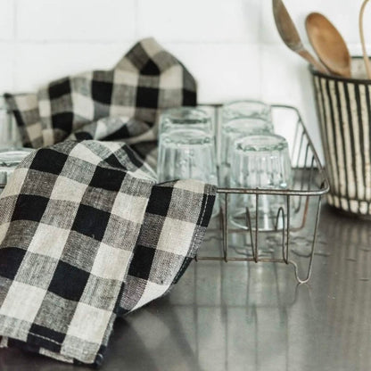 A black and beige buffalo plaid kitchen towel draped over a metal dish rack holding clear drinking glasses on a stainless steel countertop, with a striped utensil holder and wooden spoons in the background.