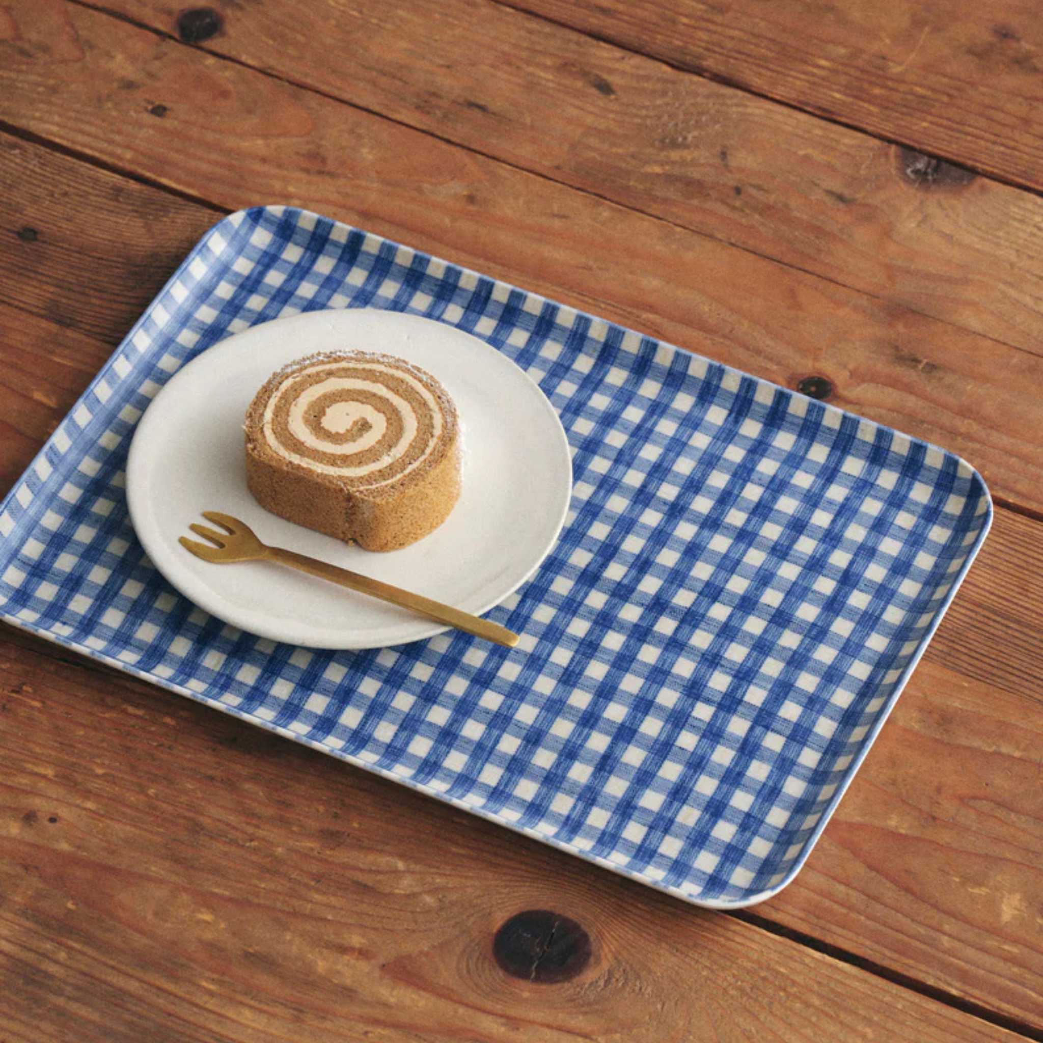 A rectangular blue and white gingham patterned coated linen tray on a wooden surface with a round plate on top containing a swiss roll cake and a gold fork.