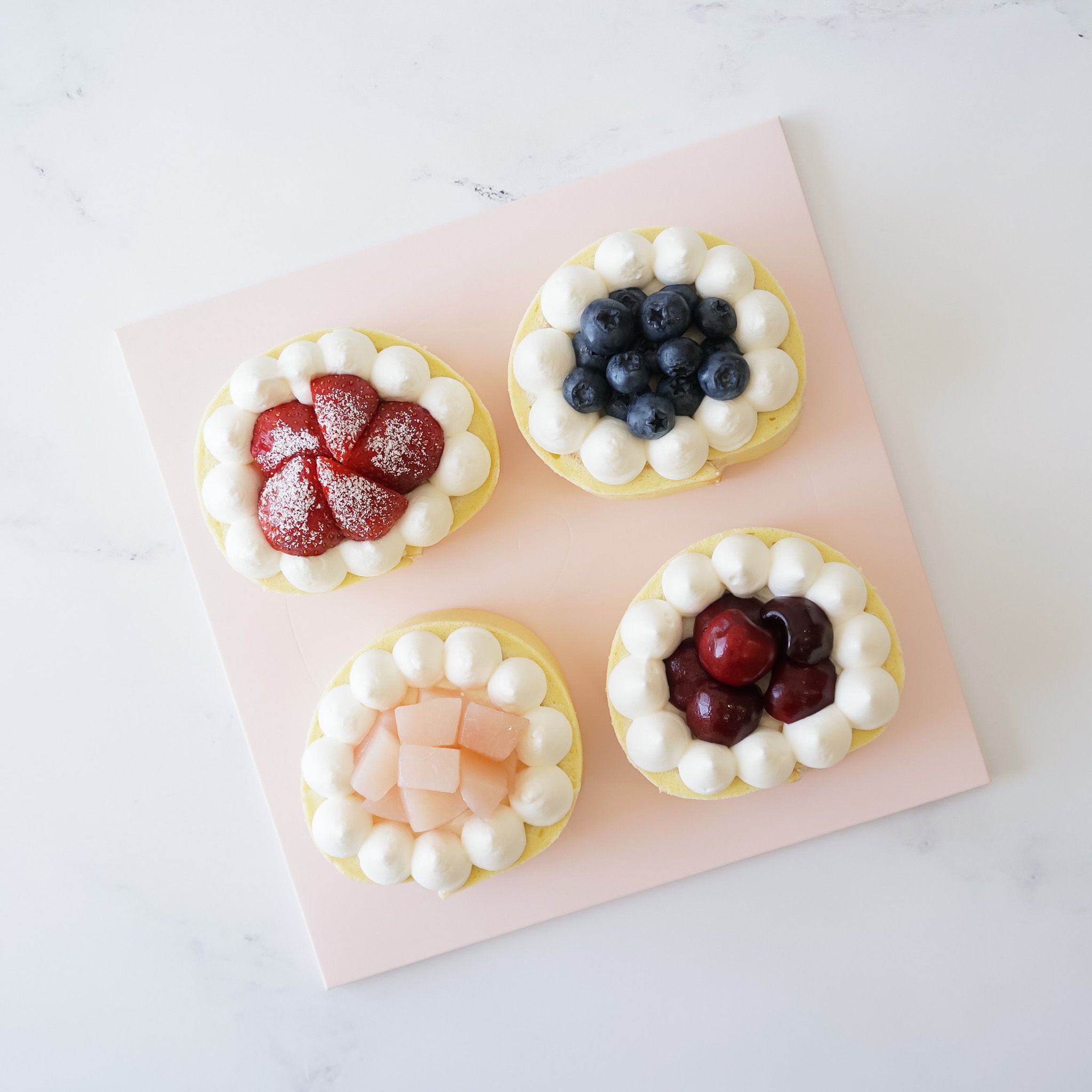 Four small round cakes with different toppings on a pink cake board. The cakes are topped with whipped cream and various fruits, including blueberries, strawberries, cherries, and peach slices.