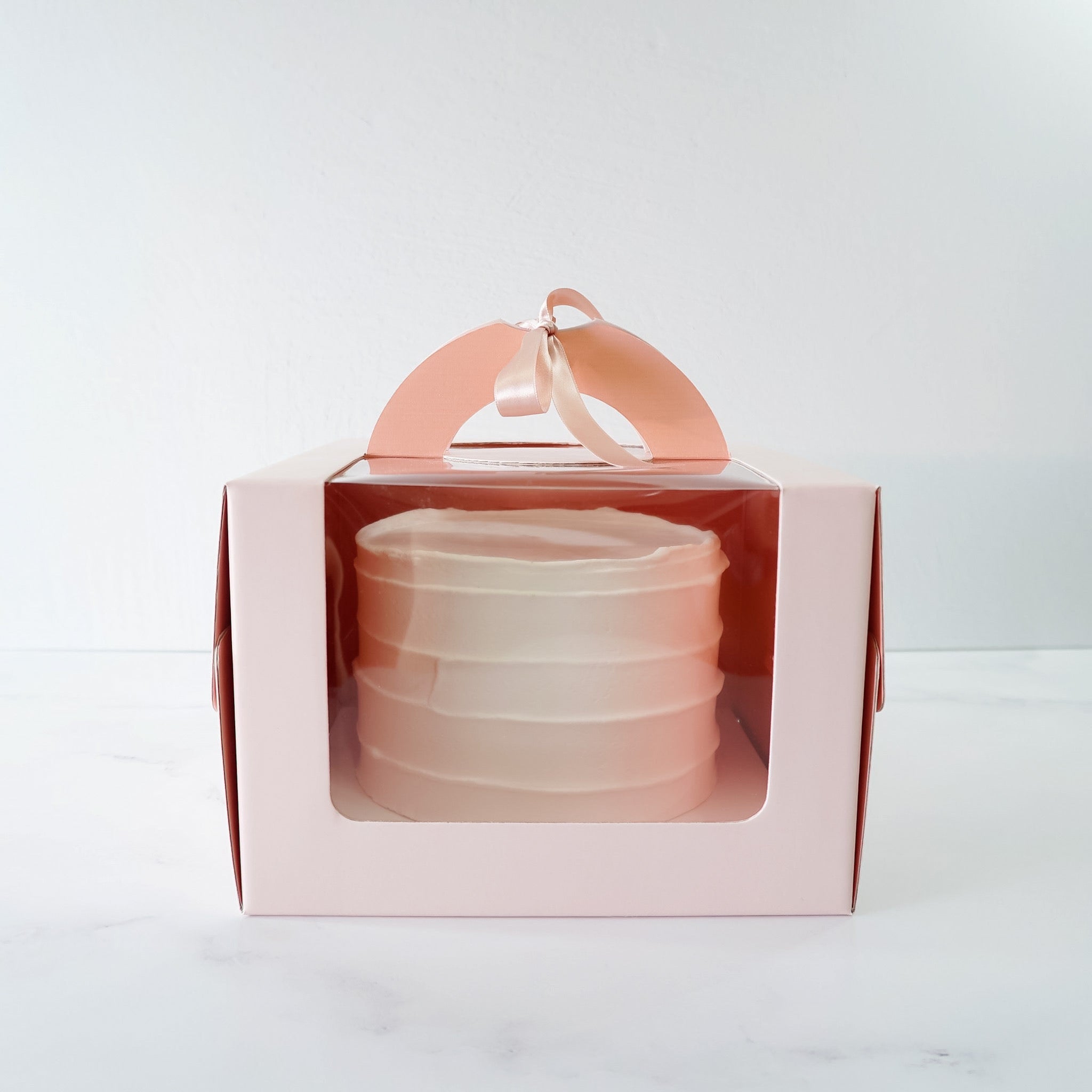 A blush pink cake box with a window, assembled and flat-packed, with a handle on top.
