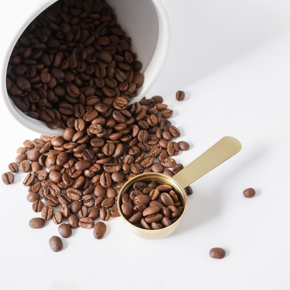 Roasted coffee beans spilling from a white ceramic container onto a white surface, with a small gold measuring cup holding some beans in the foreground.