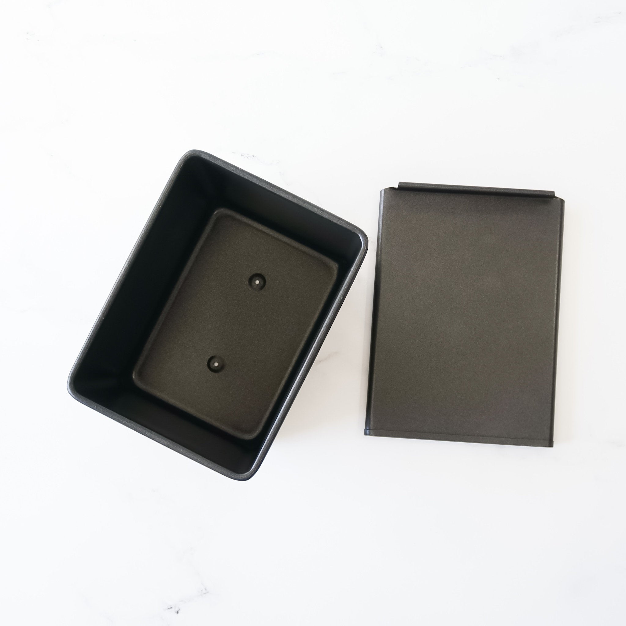 A black rectangular loaf pan with a removable lid. The pan is empty and has two small holes on the bottom.