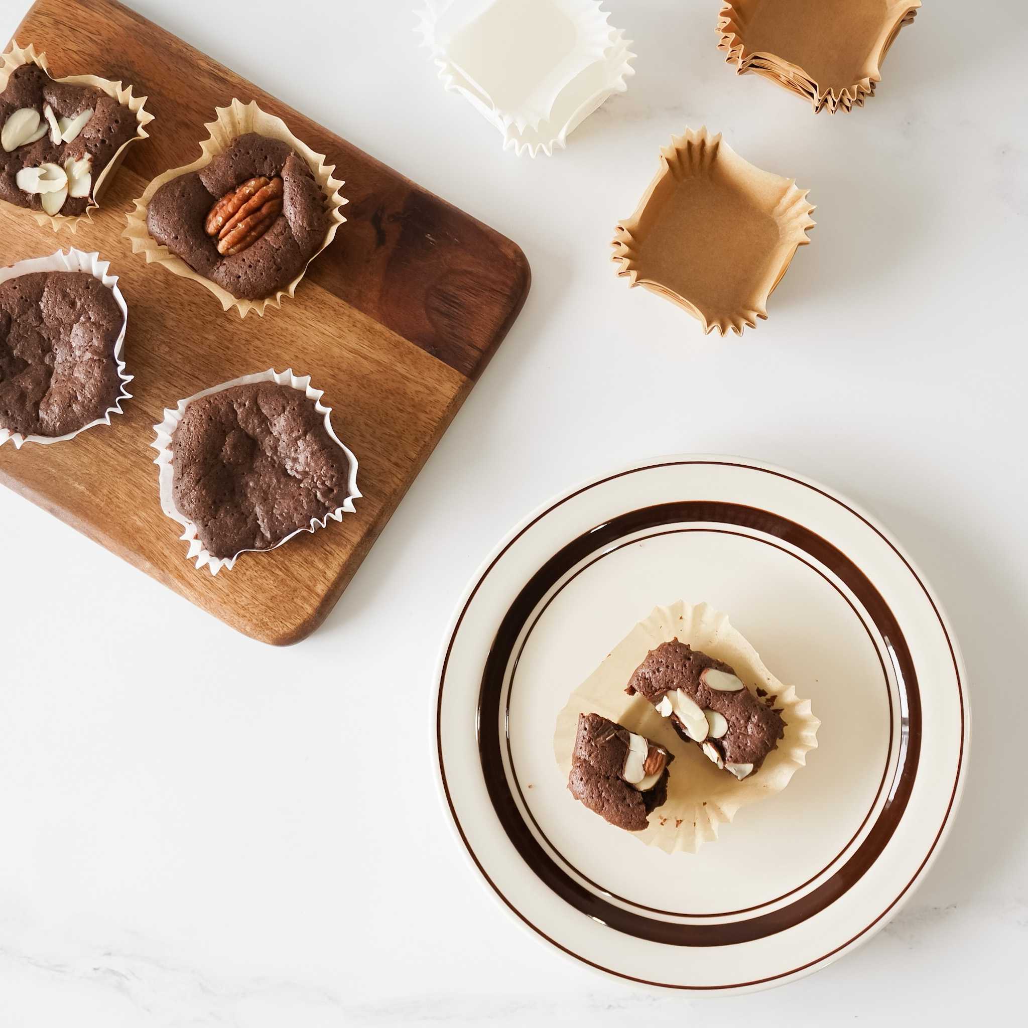 A plate with brown and white stripes holds two broken pieces of chocolate brownie in a white paper cupcake liner. The plate is on a white surface.  There are four chocolate brownies in white paper cupcake liners on a wooden cutting board.  There are also three empty paper cupcake liners on the white surface.