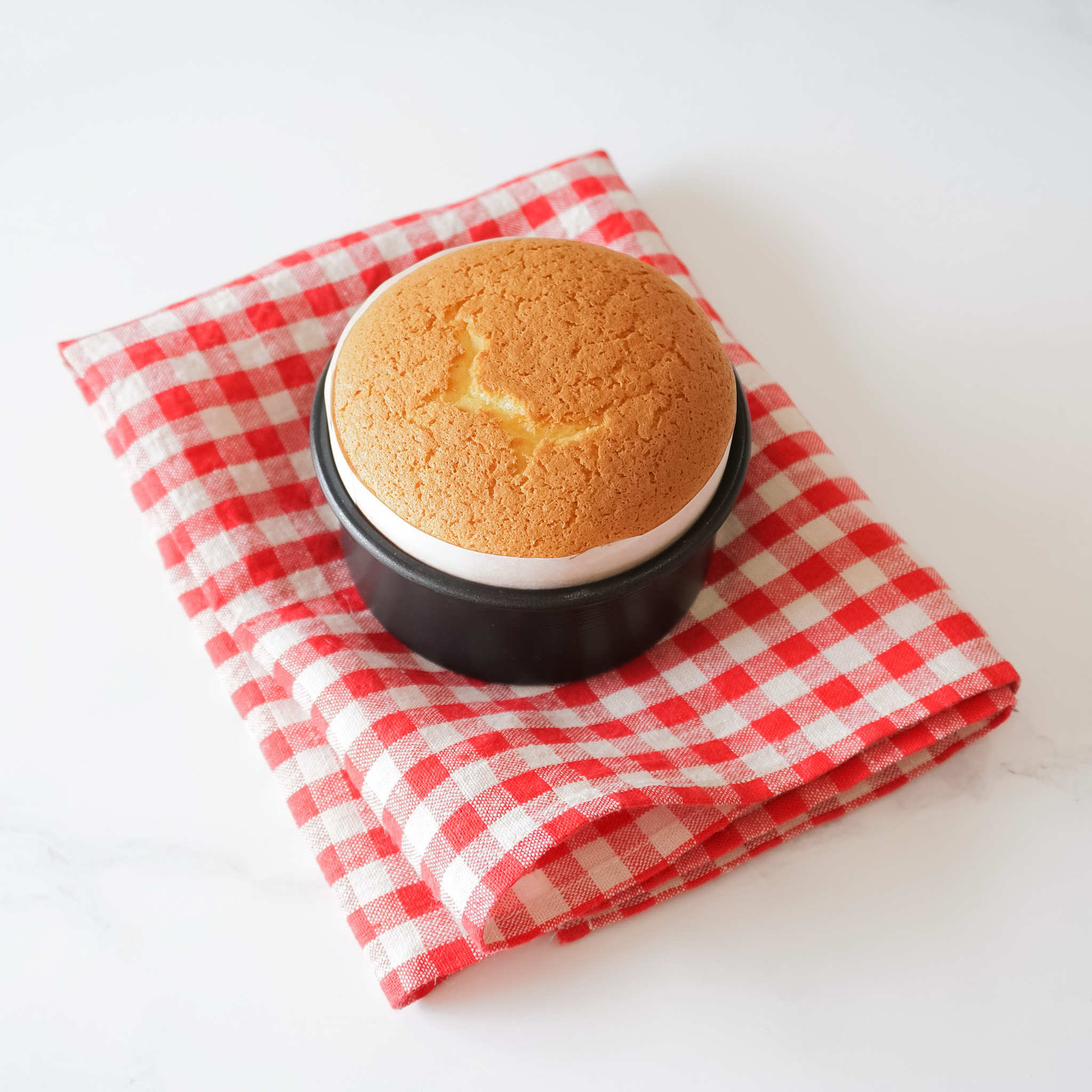 A small, freshly baked cake in a round cake pan on top of a red and white checkered kitchen cloth.