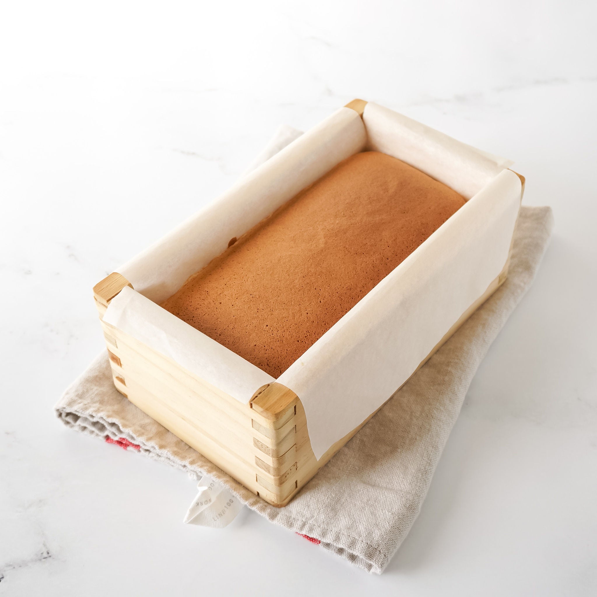 A freshly baked Castella cake in a wooden mold, which is placed on a cloth surface on a white background.