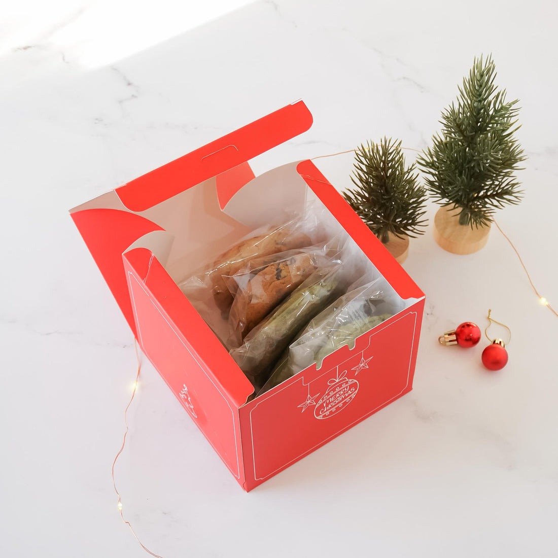 A red Christmas cookie box with a white lid is open, revealing assorted cookies in clear plastic bags. The box features a festive design with stars and the text &