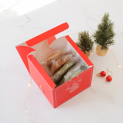 A red Christmas cookie box with a white lid is open, revealing assorted cookies in clear plastic bags. The box features a festive design with stars and the text &