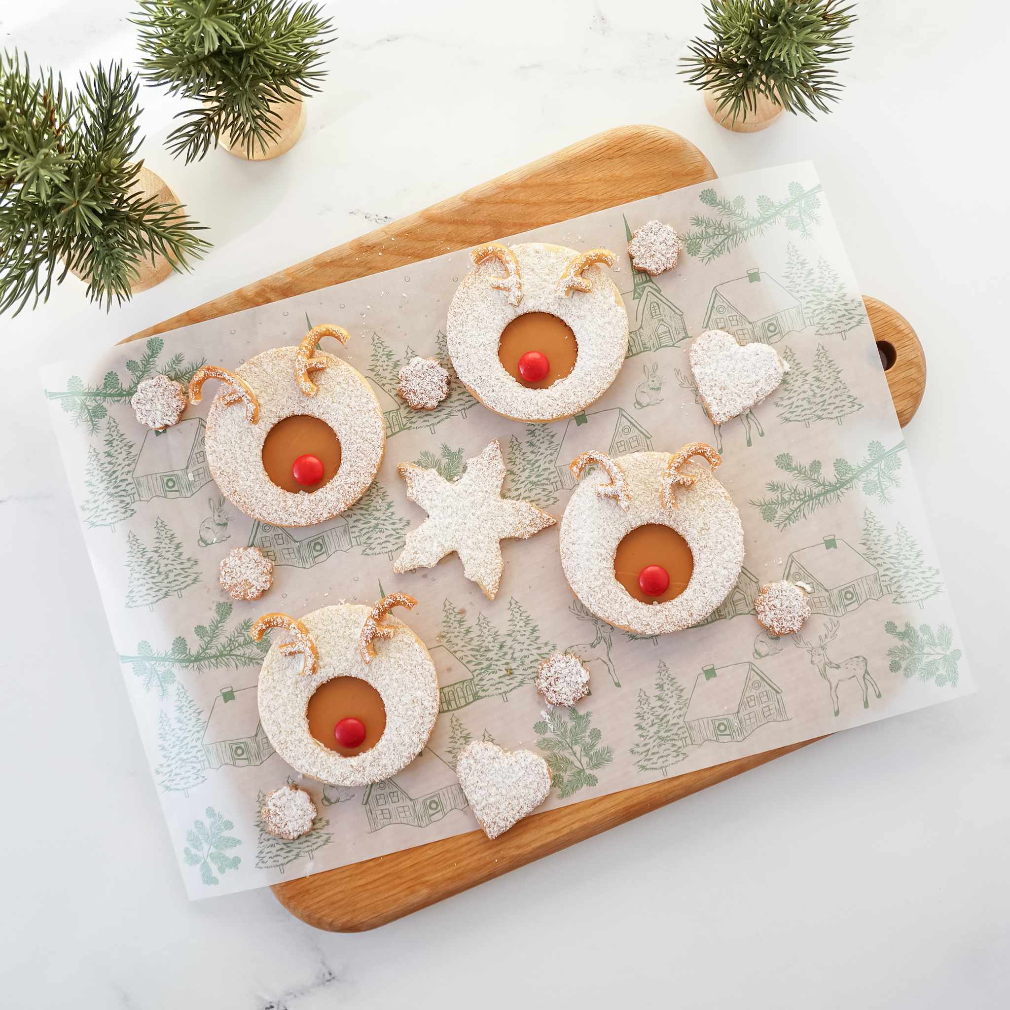 Semi-sheer deli paper with a winter village scene, including snowy houses, reindeer, rabbits, and trees, printed in classic green. The paper is displayed on a wooden board with Christmas decorations around it.