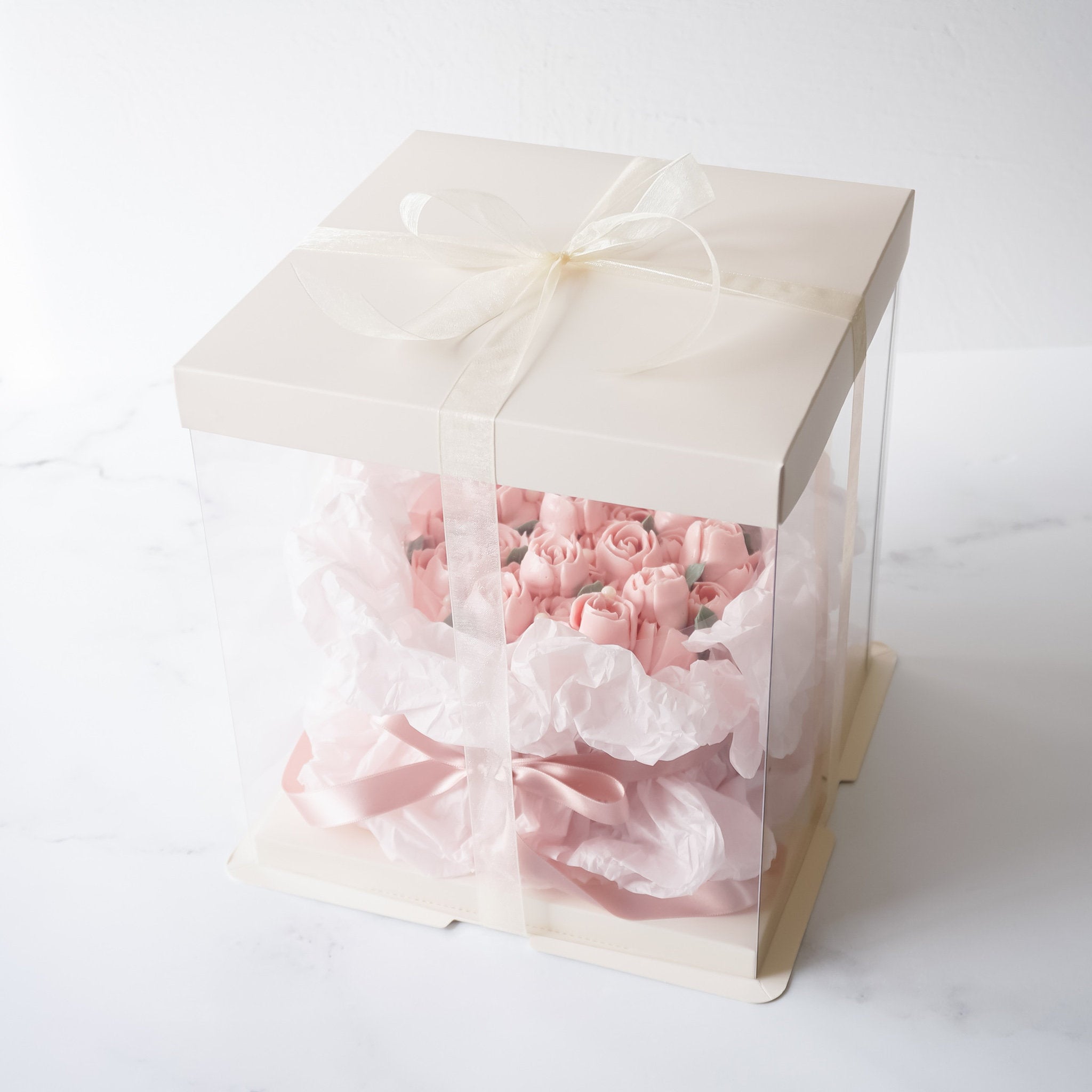 A clear plastic box with an ivory lid and a pink ribbon tied around it. Inside the box is a pink rose cake wrapped in pink tissue paper.