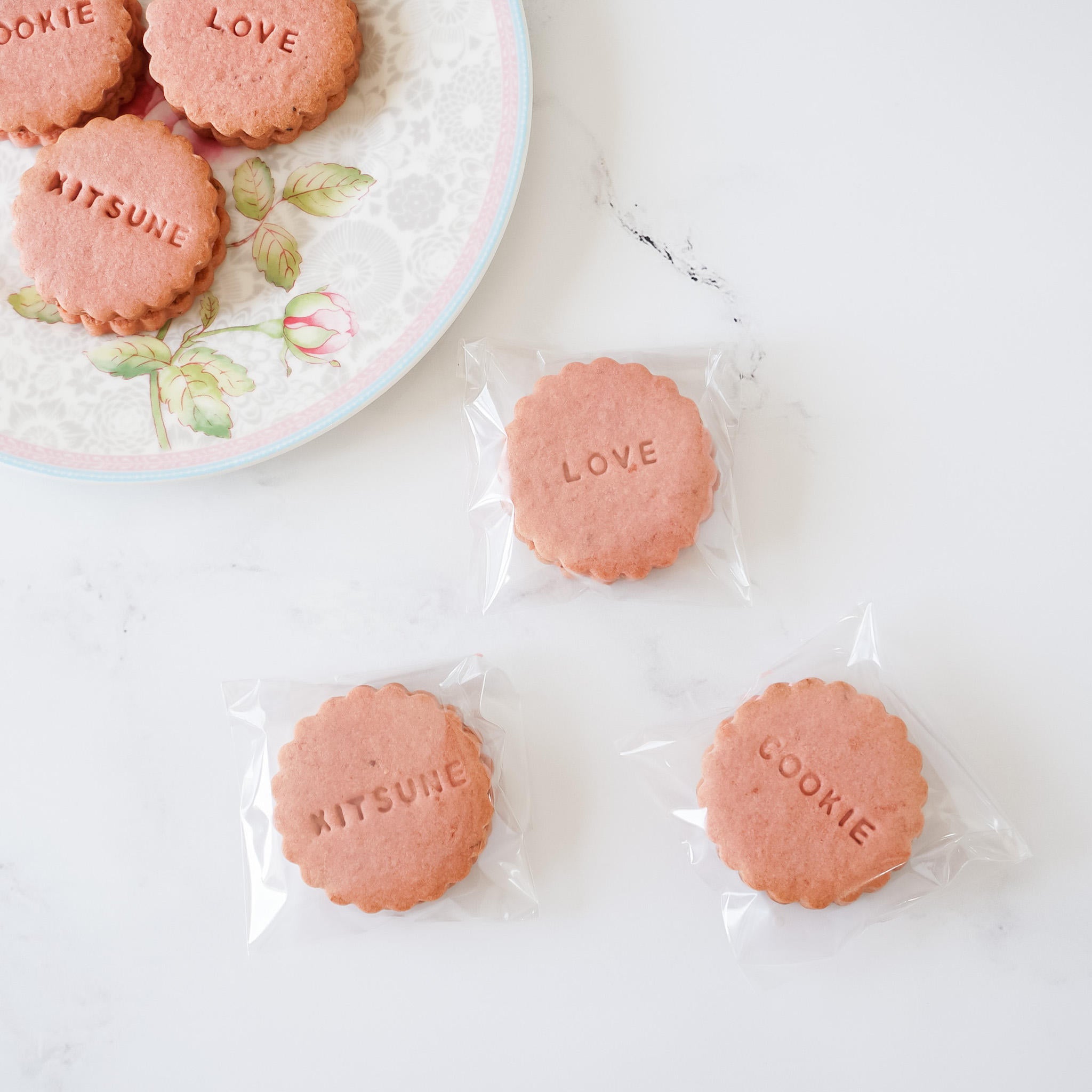 Clear cellophane bags with a pink cookie inside each, placed on a white surface, with a plate of more cookies in the background.