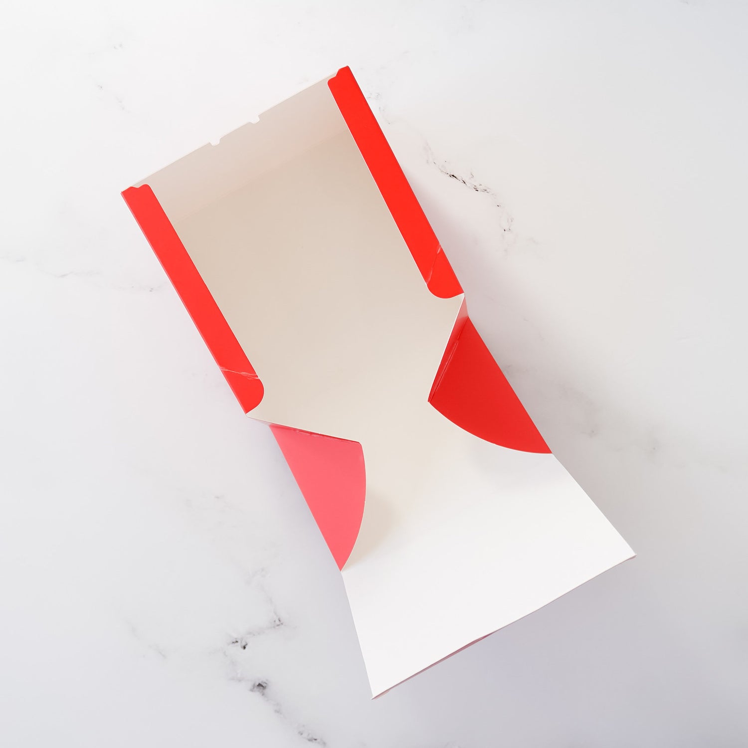 Red and white cardboard bakery box. The box is unfolded, showing its interior white surface and the red exterior. The box is designed for food packaging and has a simple, clean design.  The box is shown on a marble-like surface.
