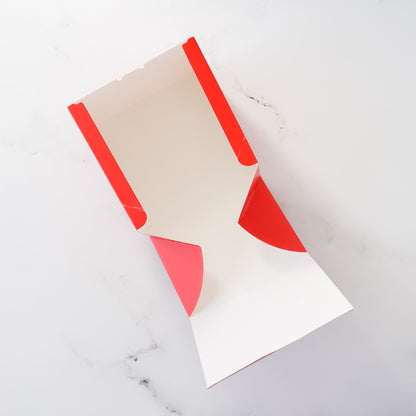 Red and white cardboard bakery box. The box is unfolded, showing its interior white surface and the red exterior. The box is designed for food packaging and has a simple, clean design.  The box is shown on a marble-like surface.