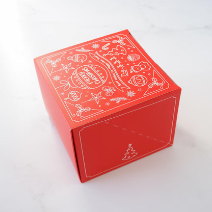 Red square Christmas gift box with white line art design. The design includes ornaments, candy canes, stars, and the text &