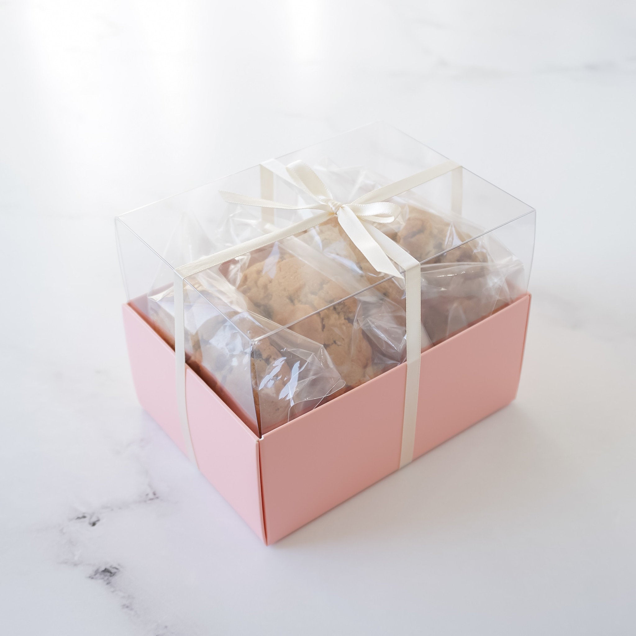 A pink gift box with a clear lid, tied with a white ribbon. The box contains multiple packages of cookies.