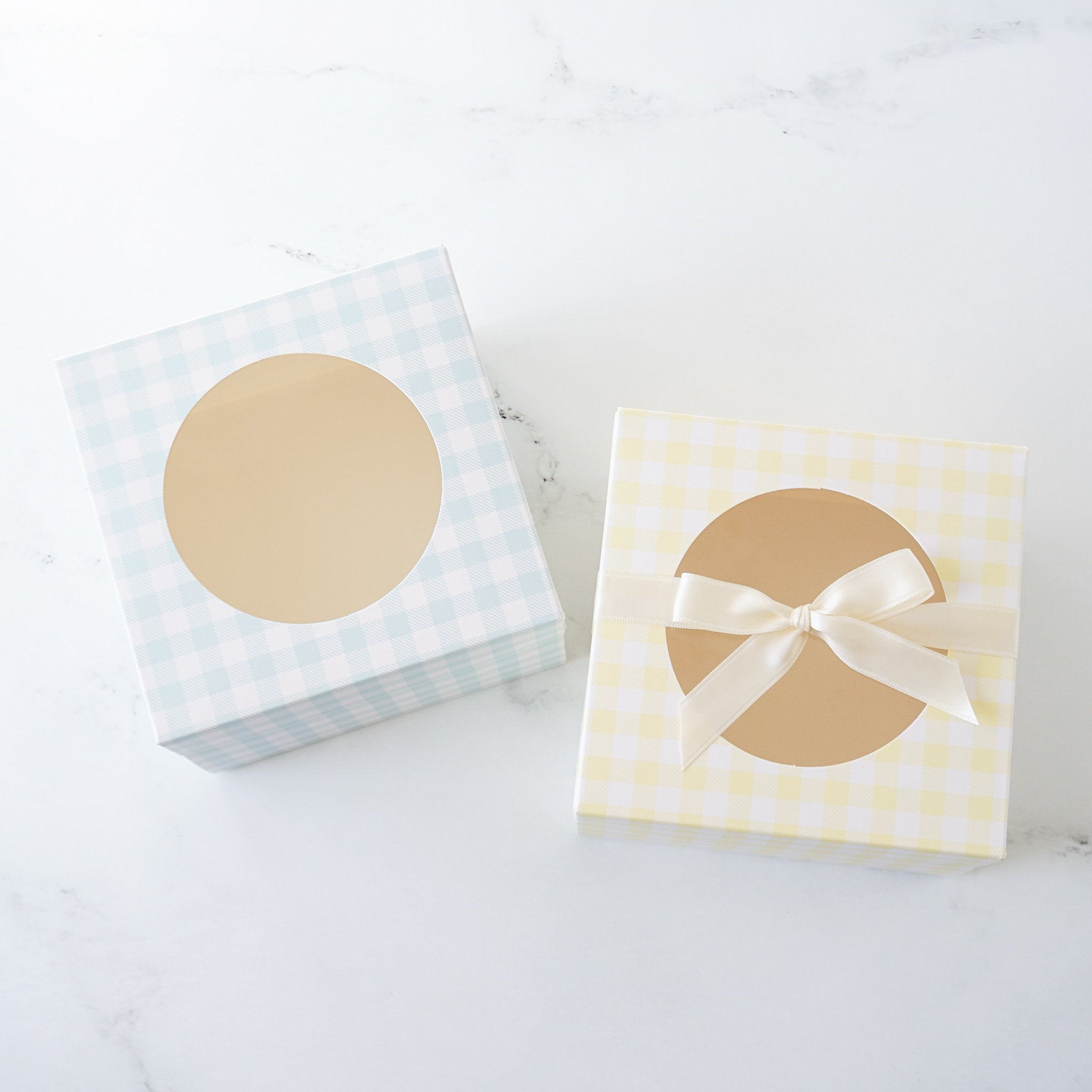 Two square gift boxes with clear windows and ribbon accents. The box on the left is light blue with a white gingham pattern. The box on the right is yellow with a white gingham pattern.