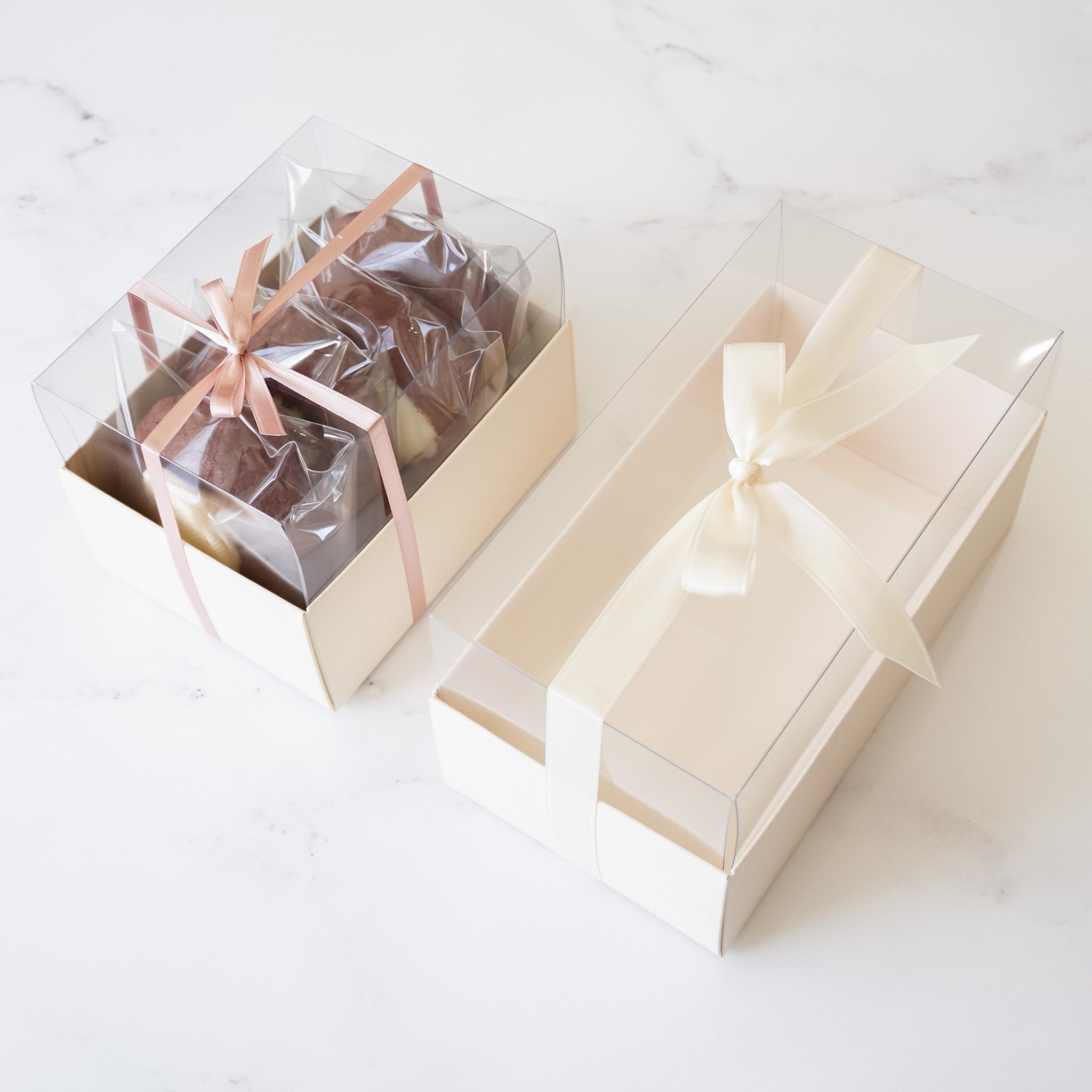 Two bakery boxes with clear lids. The boxes are ivory and have a satin ribbon tied around them. The box on the left contains cookies. The box on the right is empty.