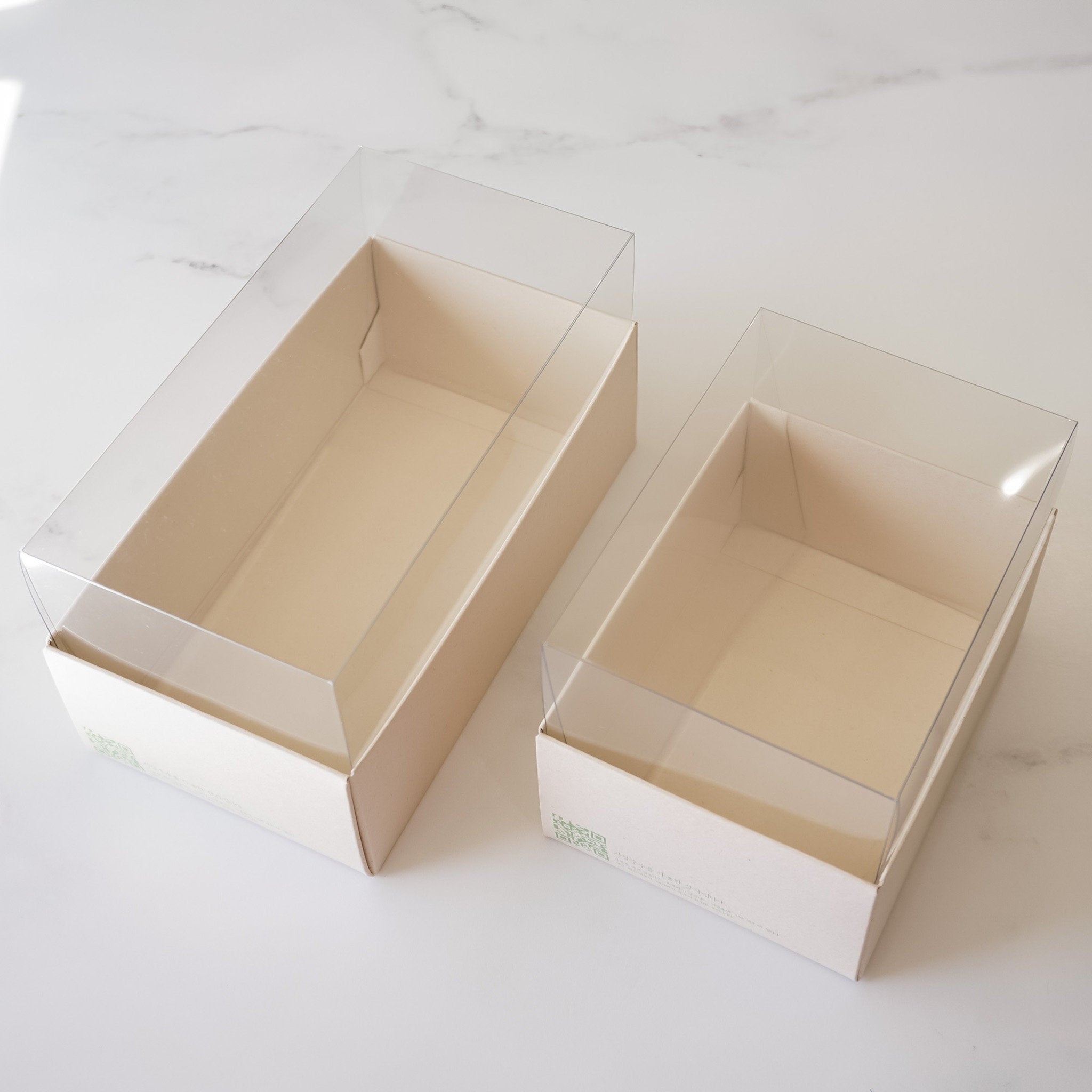 Two empty rectangular boxes with clear plastic lids. The boxes are made of light beige cardboard and are sitting on a white marble surface.