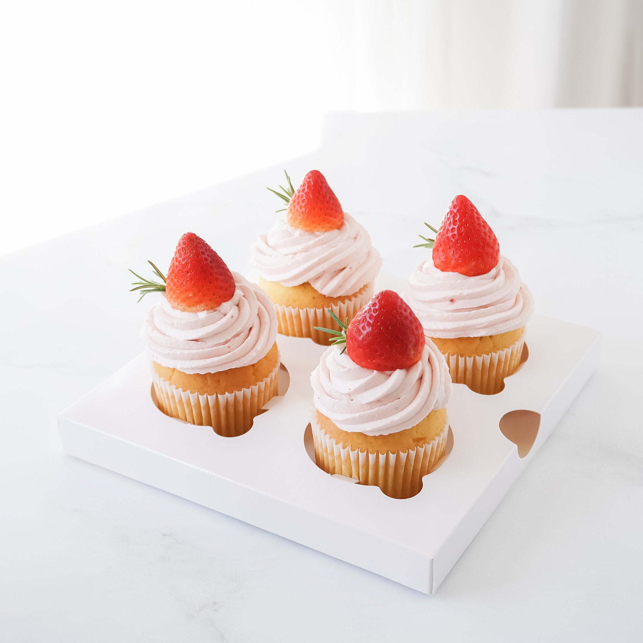 Four cupcakes with white frosting and a strawberry on top, presented in a glossy white cupcake insert.
