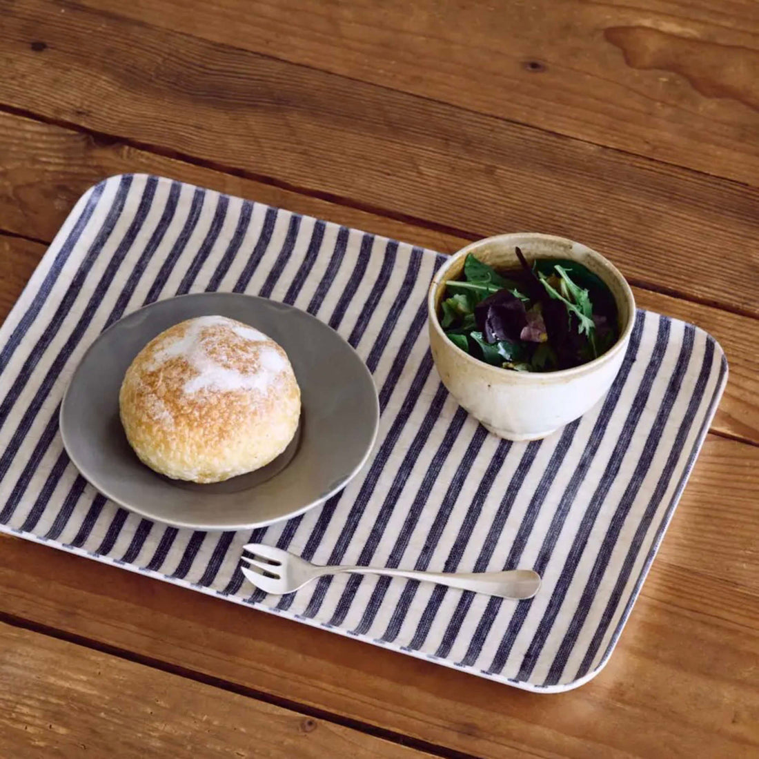 Bun on a plate with a bowl of greens on a striped tray over a wooden table
