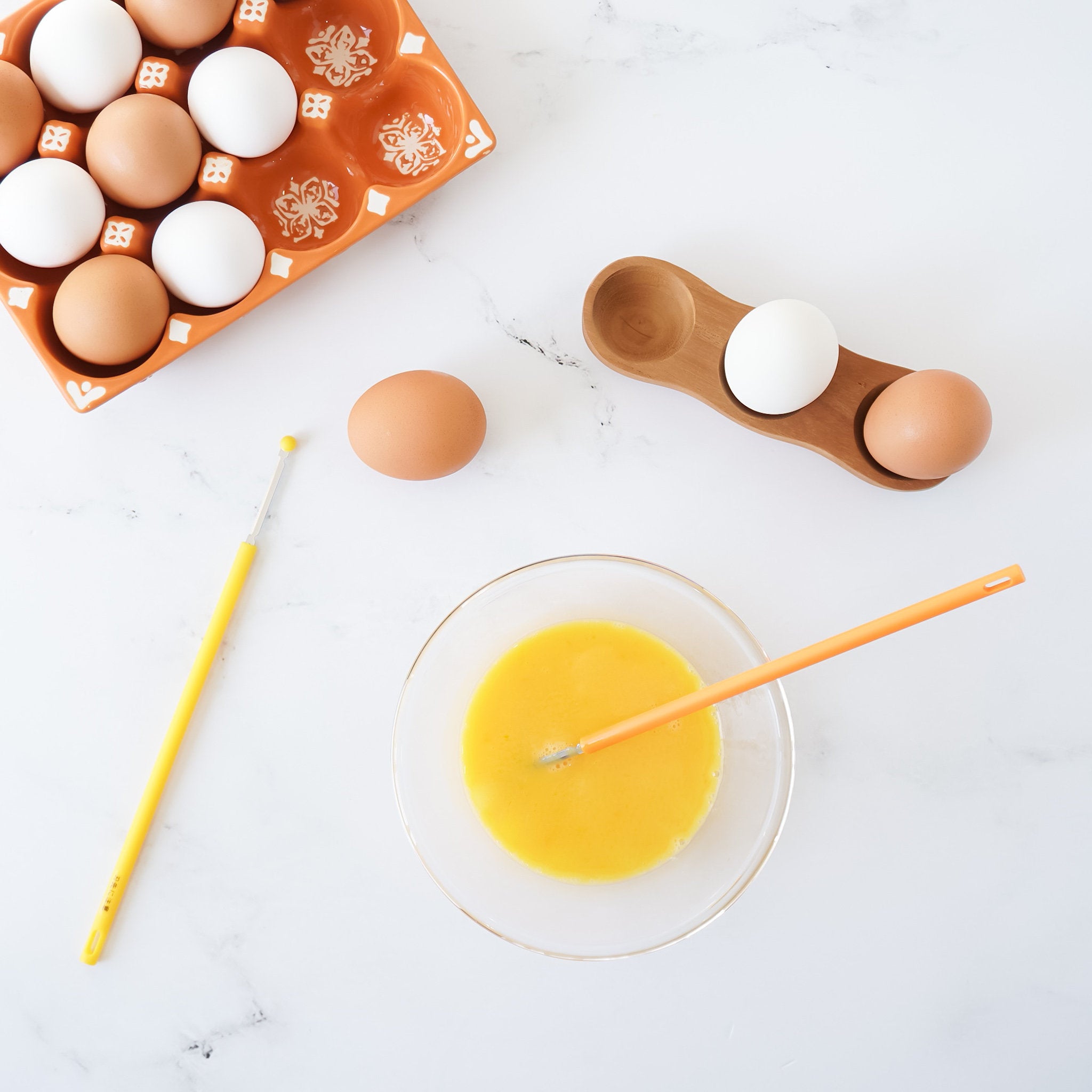 An egg whisk stick with a yellow handle and stainless steel blade is lying next to a bowl of whisked eggs, with whole eggs and a carton in the background.