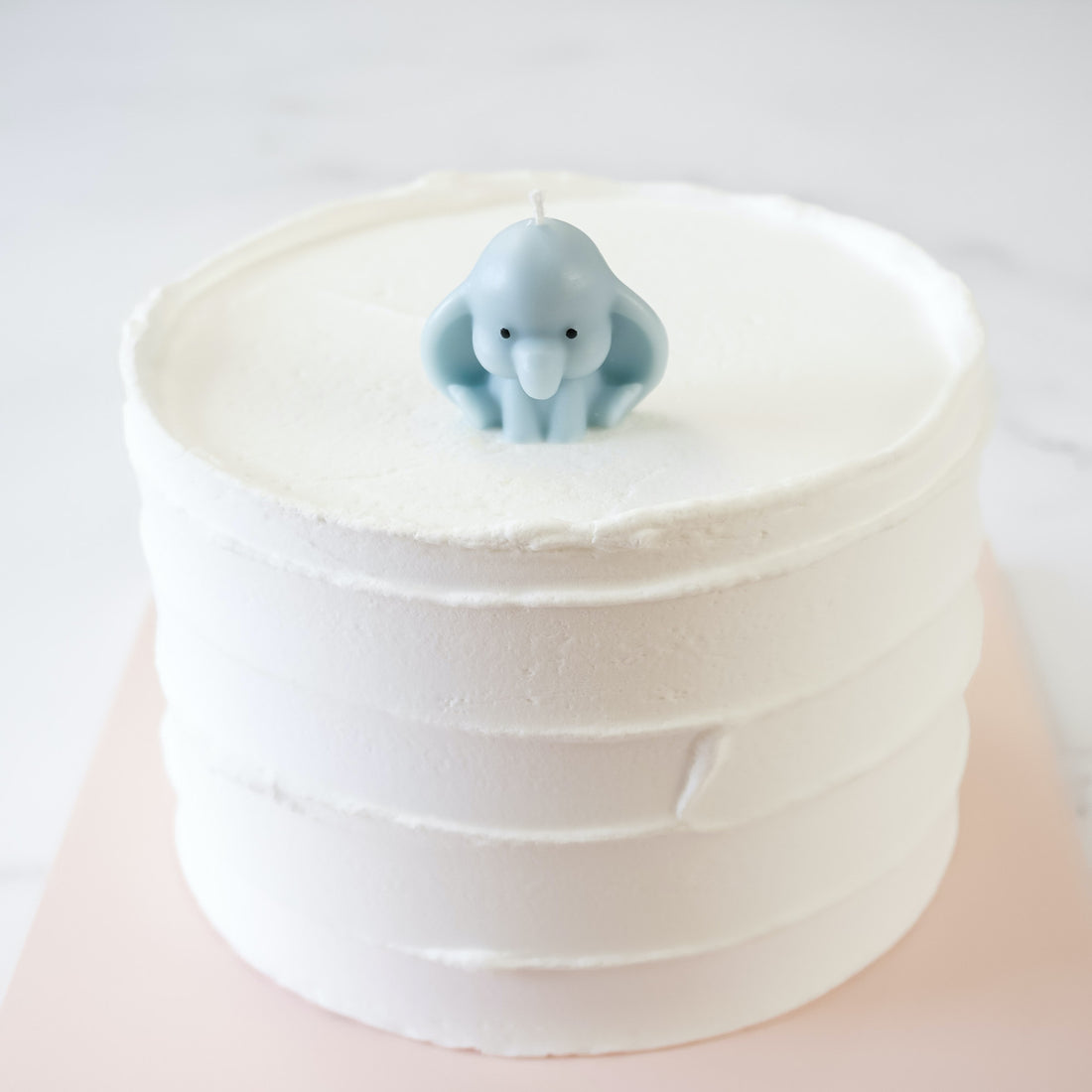 A stack of white cake layers with a small blue birthday candle in the shape of a baby elephant on top.