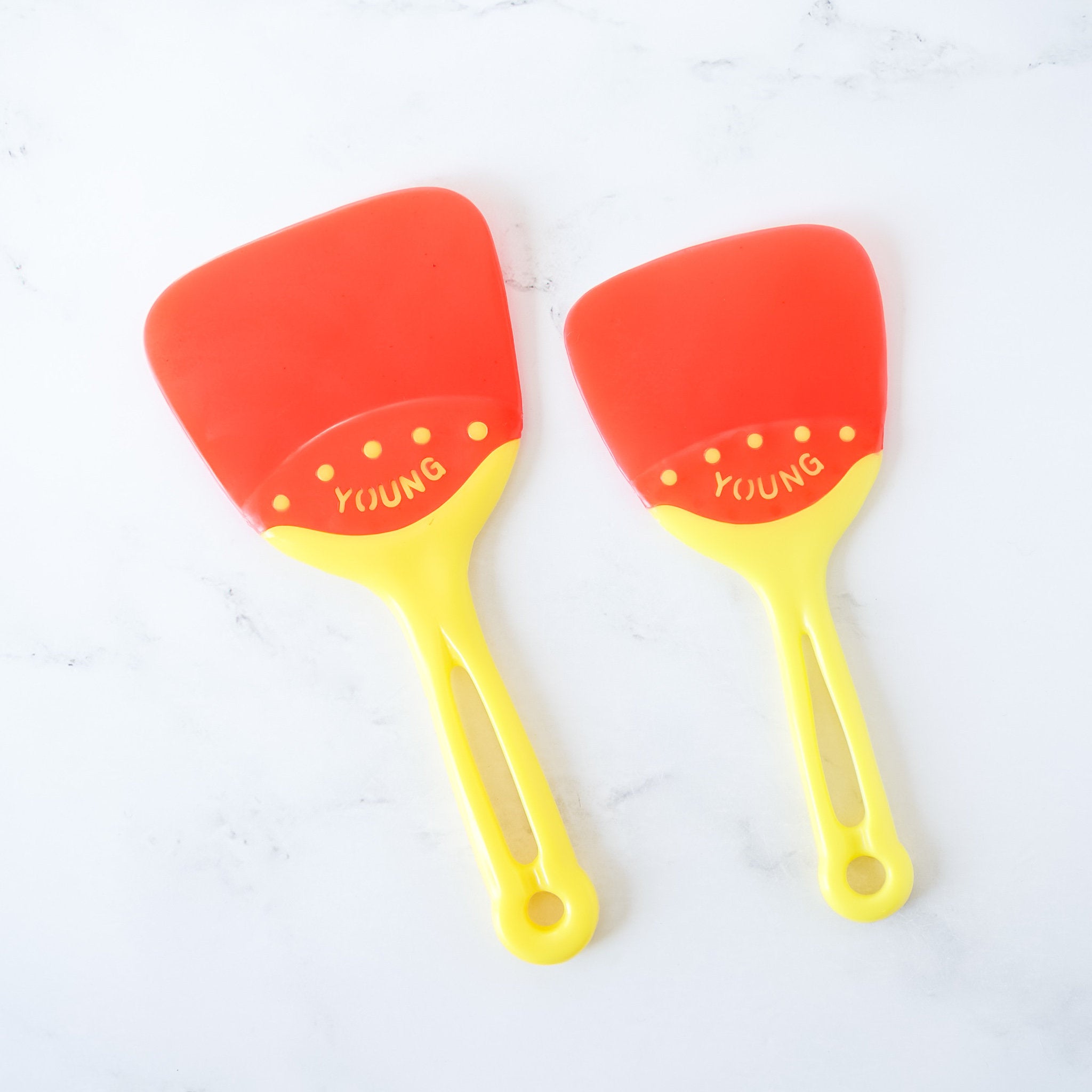 Two flexible baking spatulas with red heads and yellow handles, one large and one extra large, laid out on a white surface.