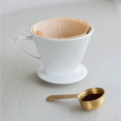 A white ceramic pour-over coffee dripper with a beige paper filter, shown on a light gray surface next to a small gold measuring spoon filled with ground coffee.