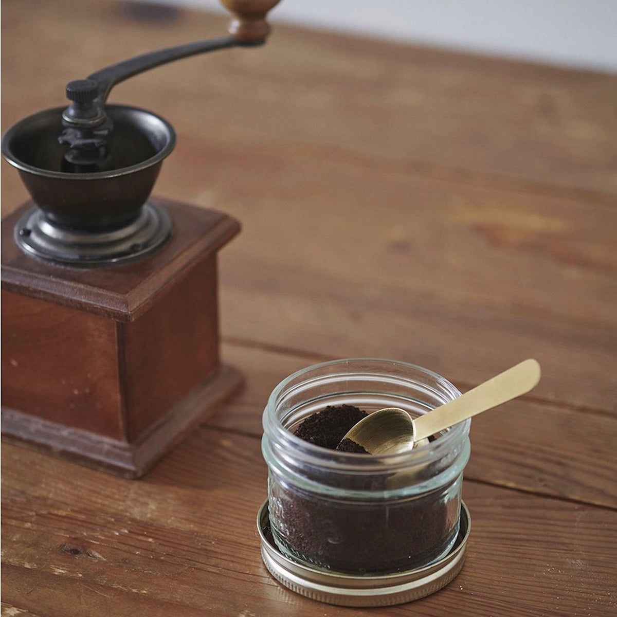 A glass jar filled with dark ground coffee sits on a wooden table, with a small gold spoon resting inside. A vintage-style manual coffee grinder is blurred in the background on the left.