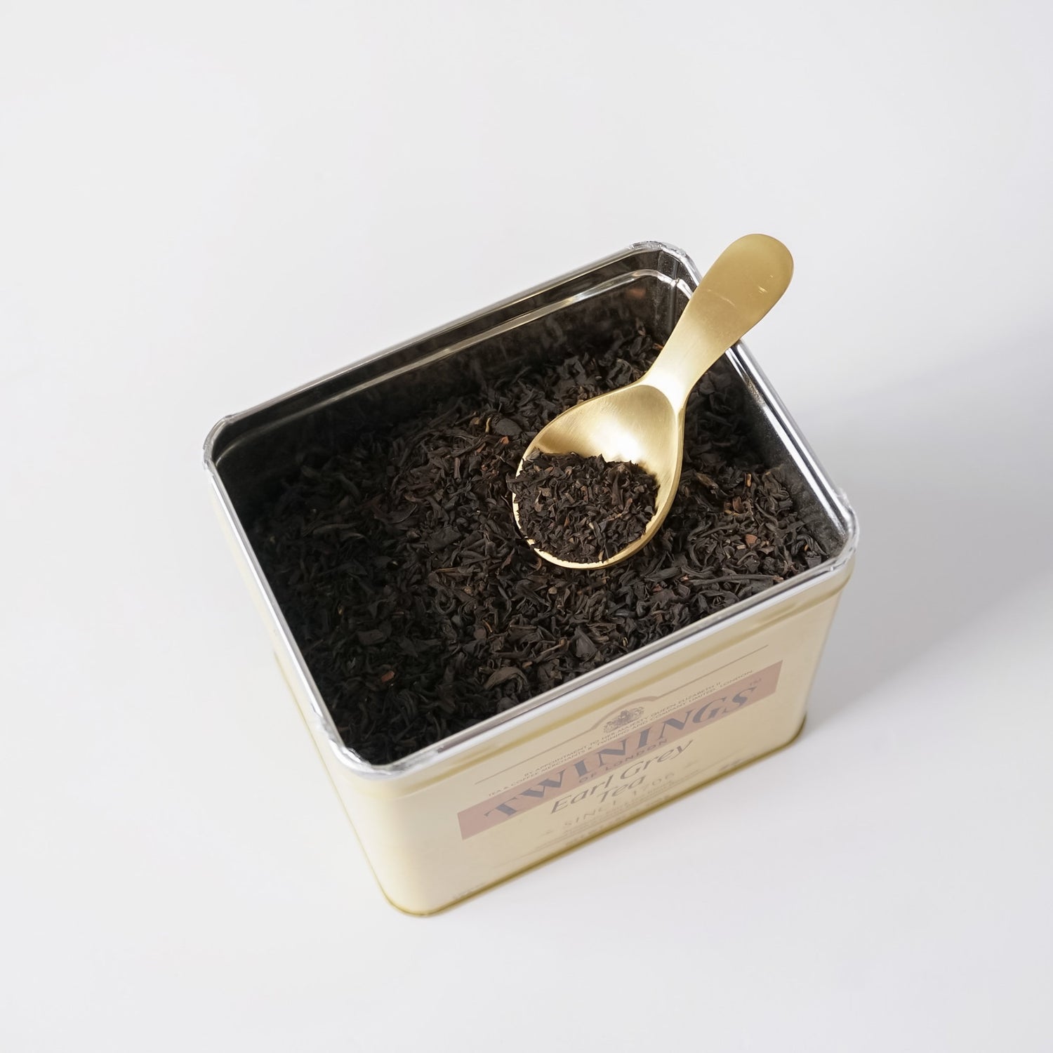 An open cream-colored metal tea tin filled with dark loose-leaf tea, with a small gold spoon resting inside. The front of the tin shows the Twinings brand name and &