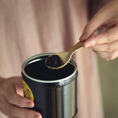 A close-up lifestyle shot of a person holding a black metal tin of loose leaf tea while using a small brass spoon to scoop the dark tea leaves.