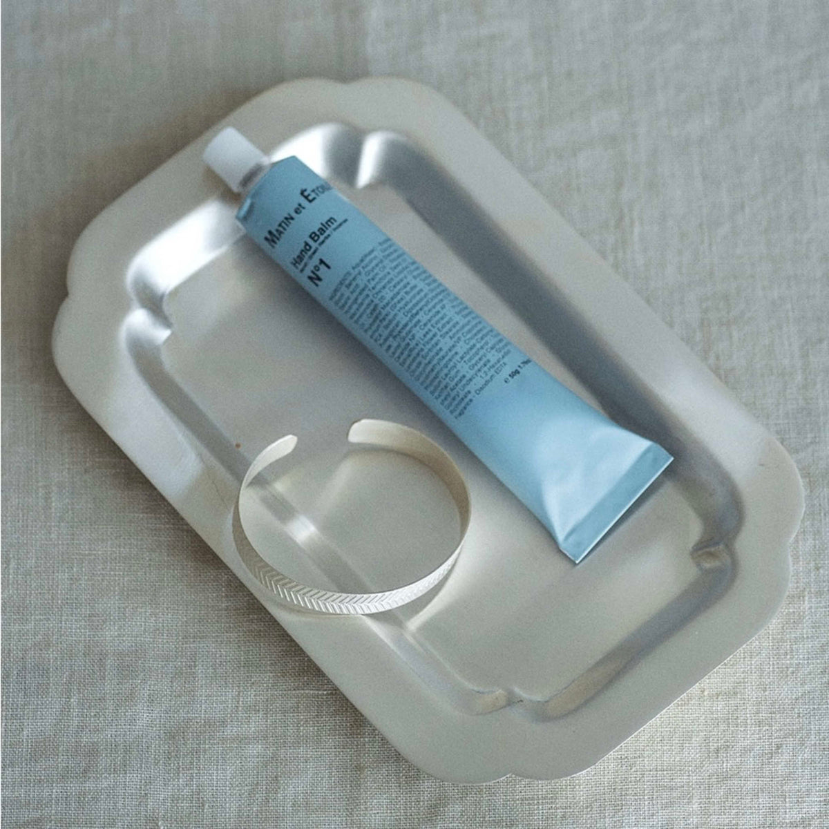 A light blue tube of hand balm rests inside a white scalloped metal dish on a textured beige linen background.